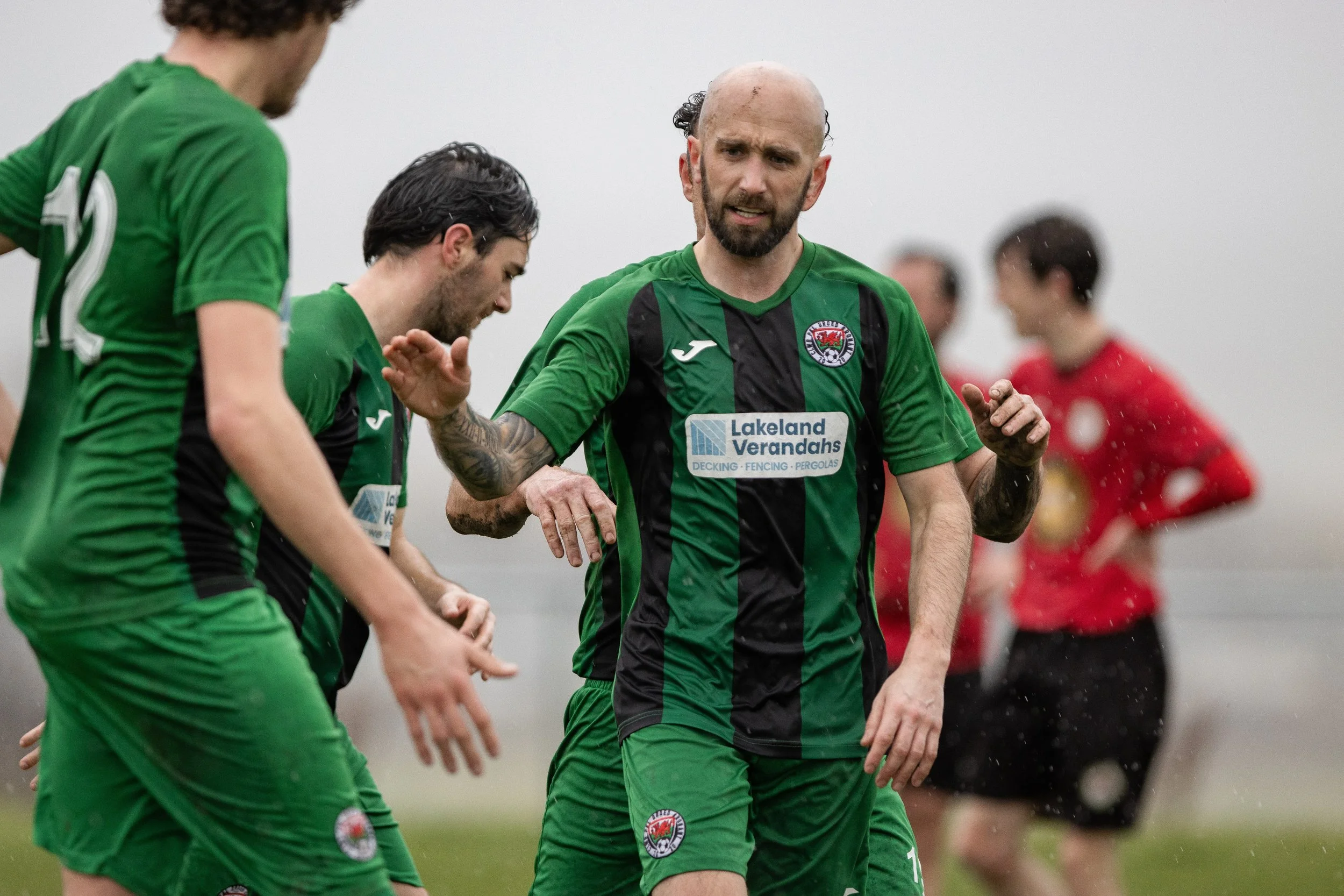 Soccer players in green and black uniforms on a field, with one player prominently in the center and a referee in a red shirt in the background.