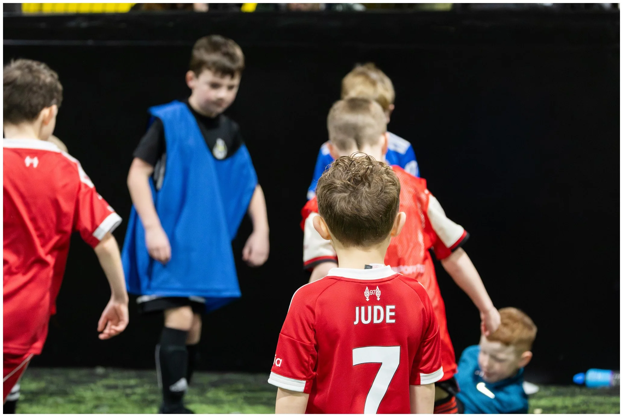 Young children playing indoor soccer. A boy with a red jersey marked "JUDE" and the number 7 is in the foreground. Other children in red, blue, and black jerseys are in the background, some standing and others sitting or kneeling on the turf.