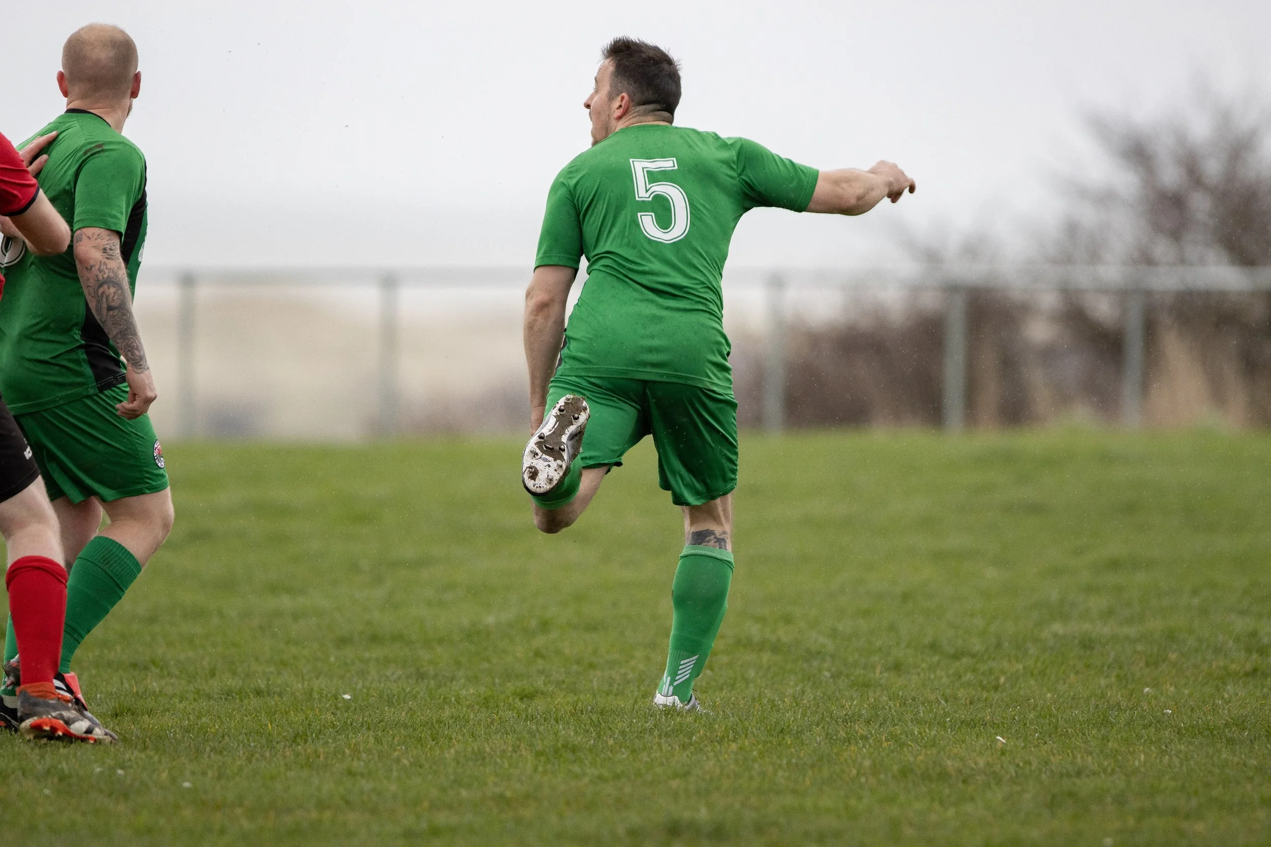 Soccer player wearing green jersey with the number 5 on the back, kicking a soccer ball during a game on a grassy field.
