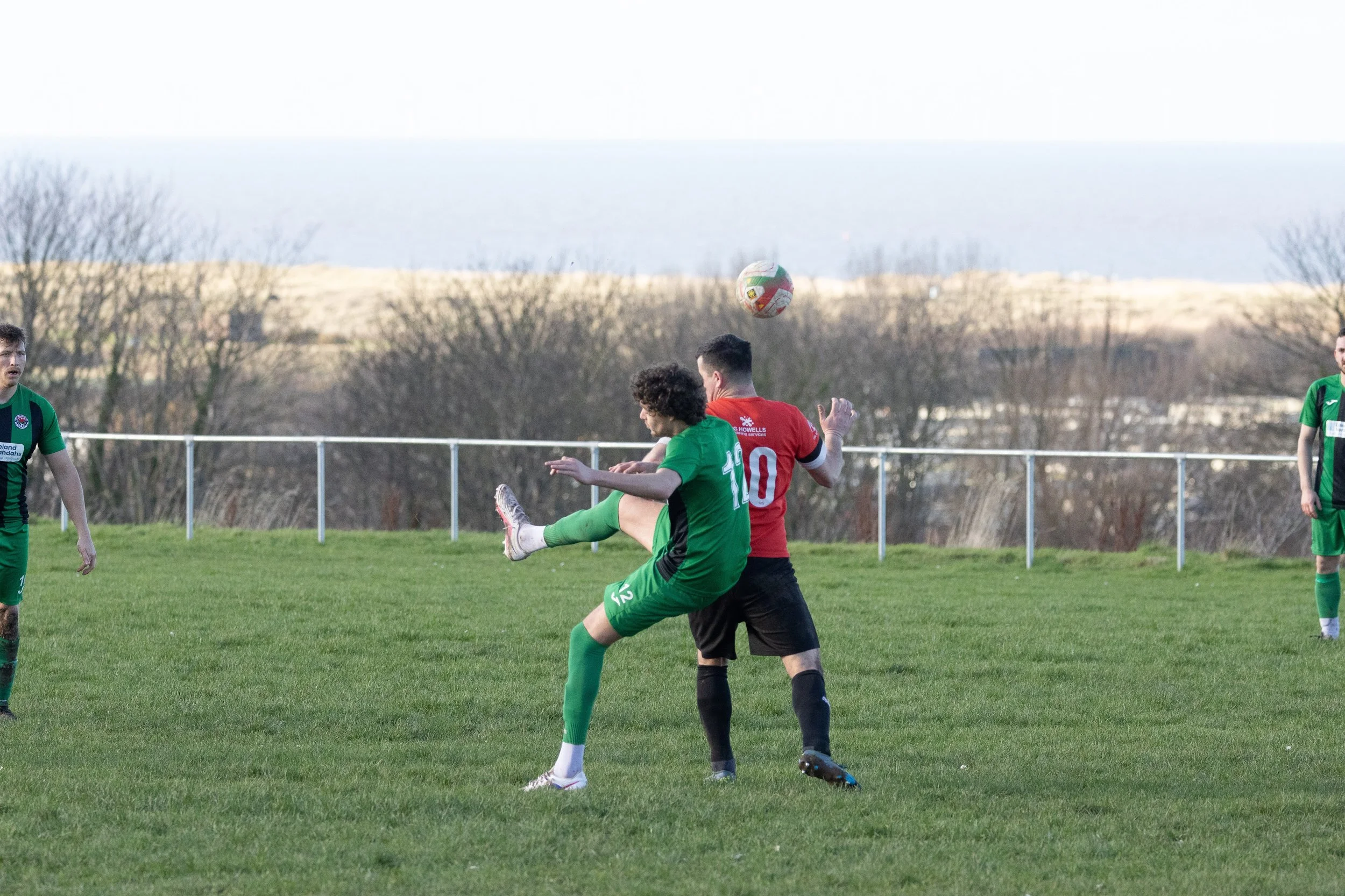 Soccer players in green and red jerseys competing for the ball on a grassy field with trees and water in the background.