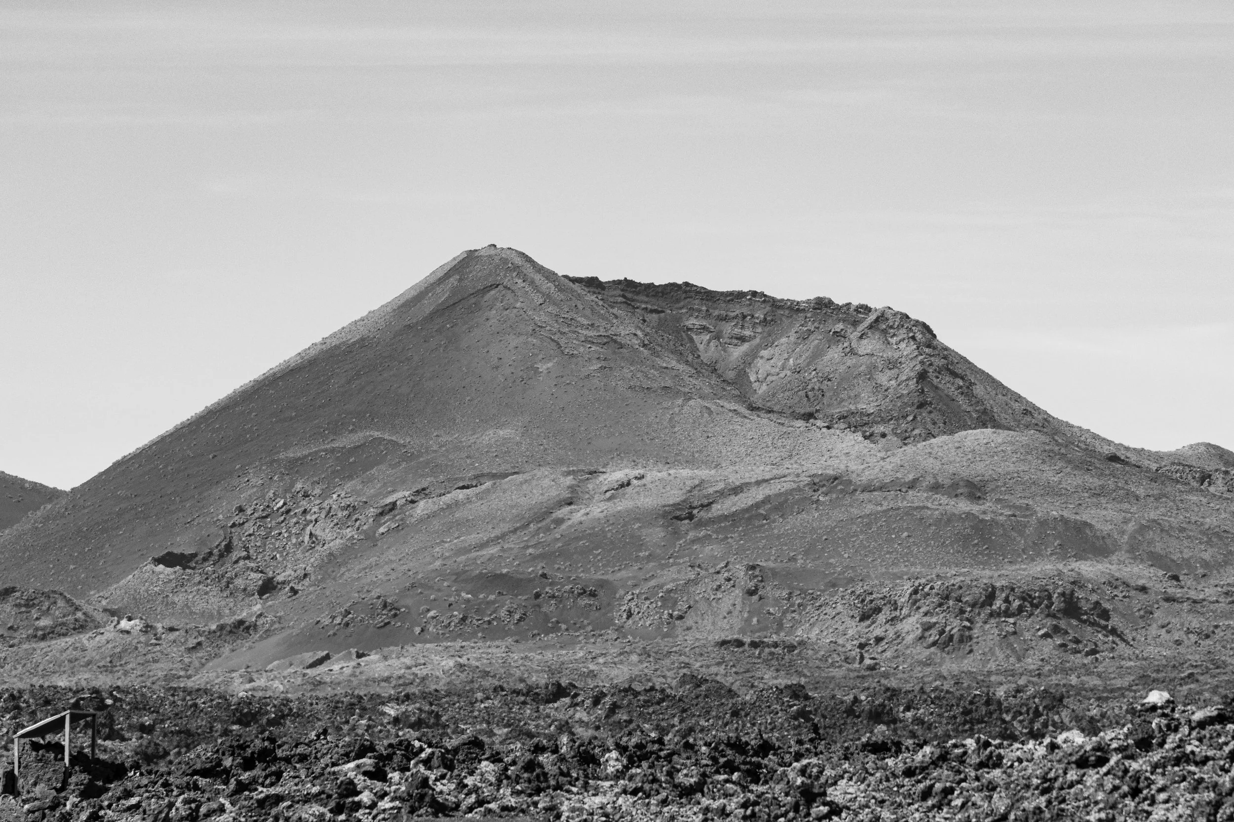 Black and white image of a volcanic mountain with a slope leading to a crater at the top and rocky terrain in the foreground.