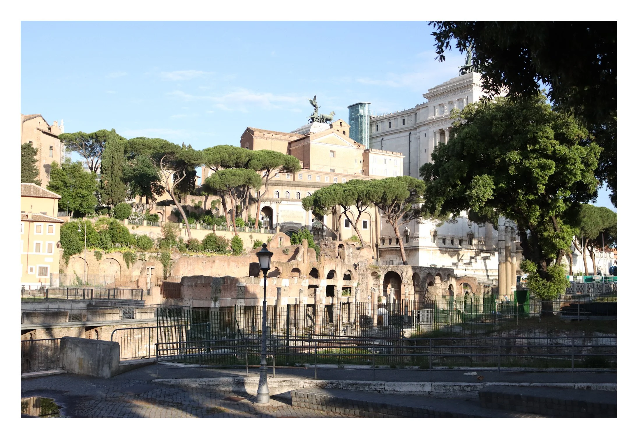 Ancient ruins with arches and columns, surrounded by greenery including tall trees, with a large historical building and sculpture on a pedestal in the background, under a clear blue sky.