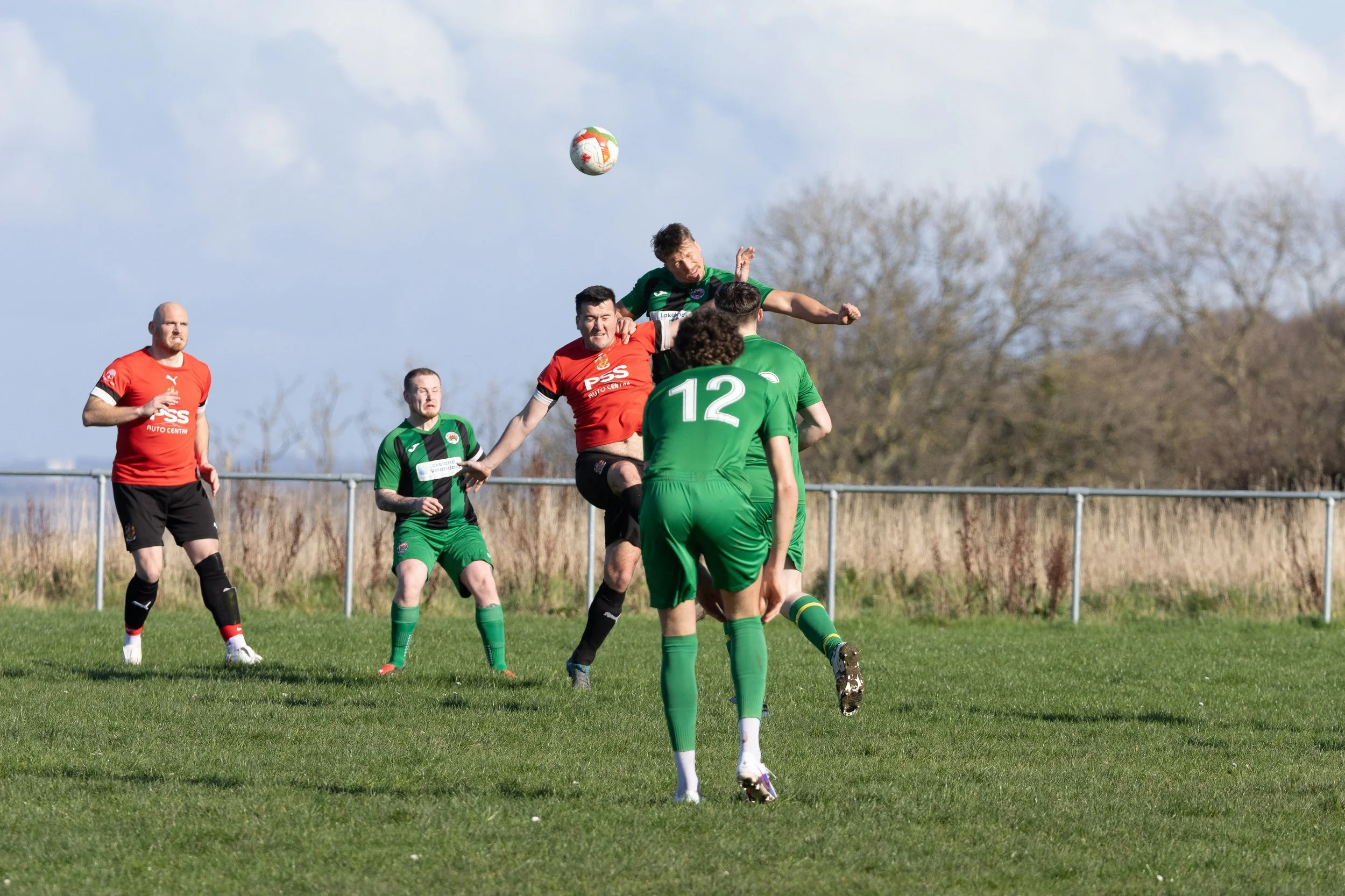 Soccer players in green and red jerseys competing for the ball during a match on a grassy field, with trees and a blue sky in the background.