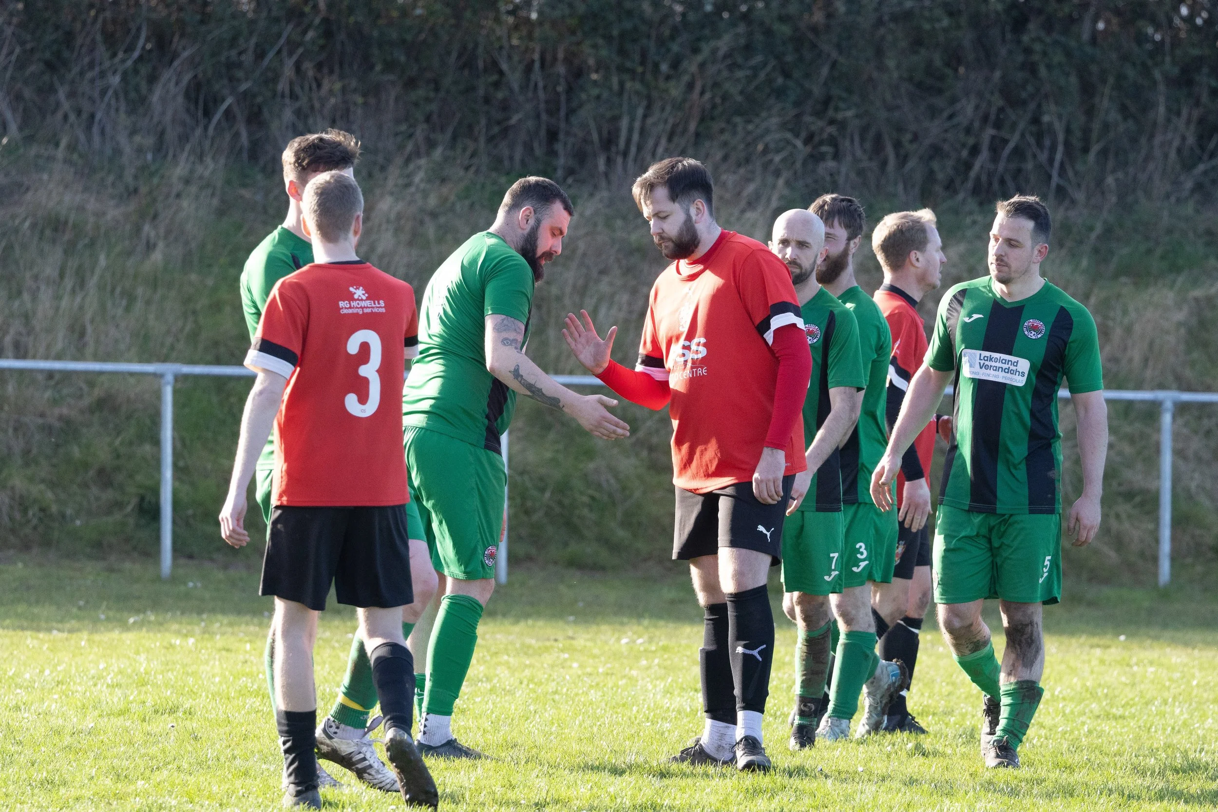 Soccer players on a field shaking hands before a match, wearing green and red jerseys.