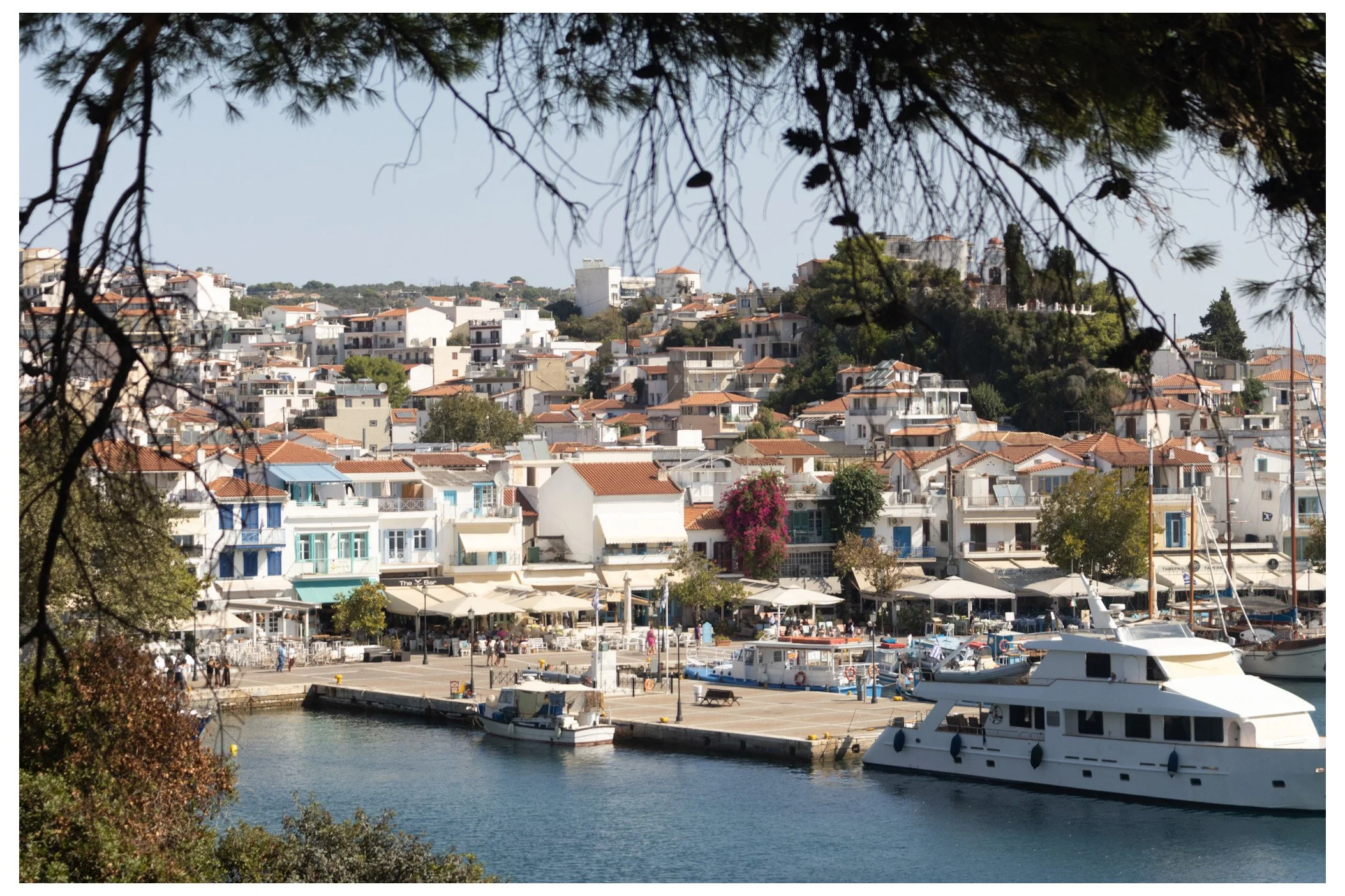 Harbor with boats docked, waterfront cafes with umbrellas, and a hillside town with white buildings and red-tiled roofs, under a clear sky.