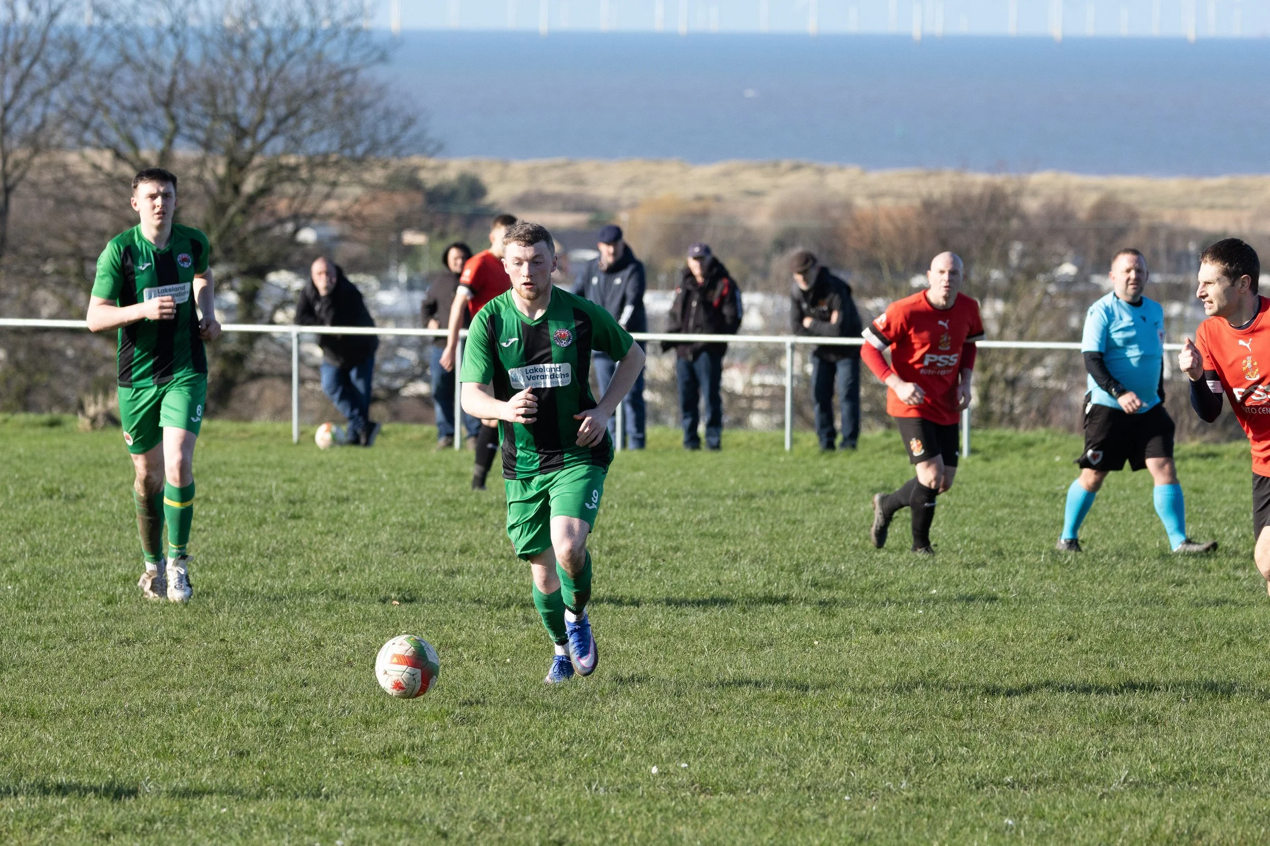 Soccer players on a grassy field during a game, with players in red and green uniforms and spectators in the background.