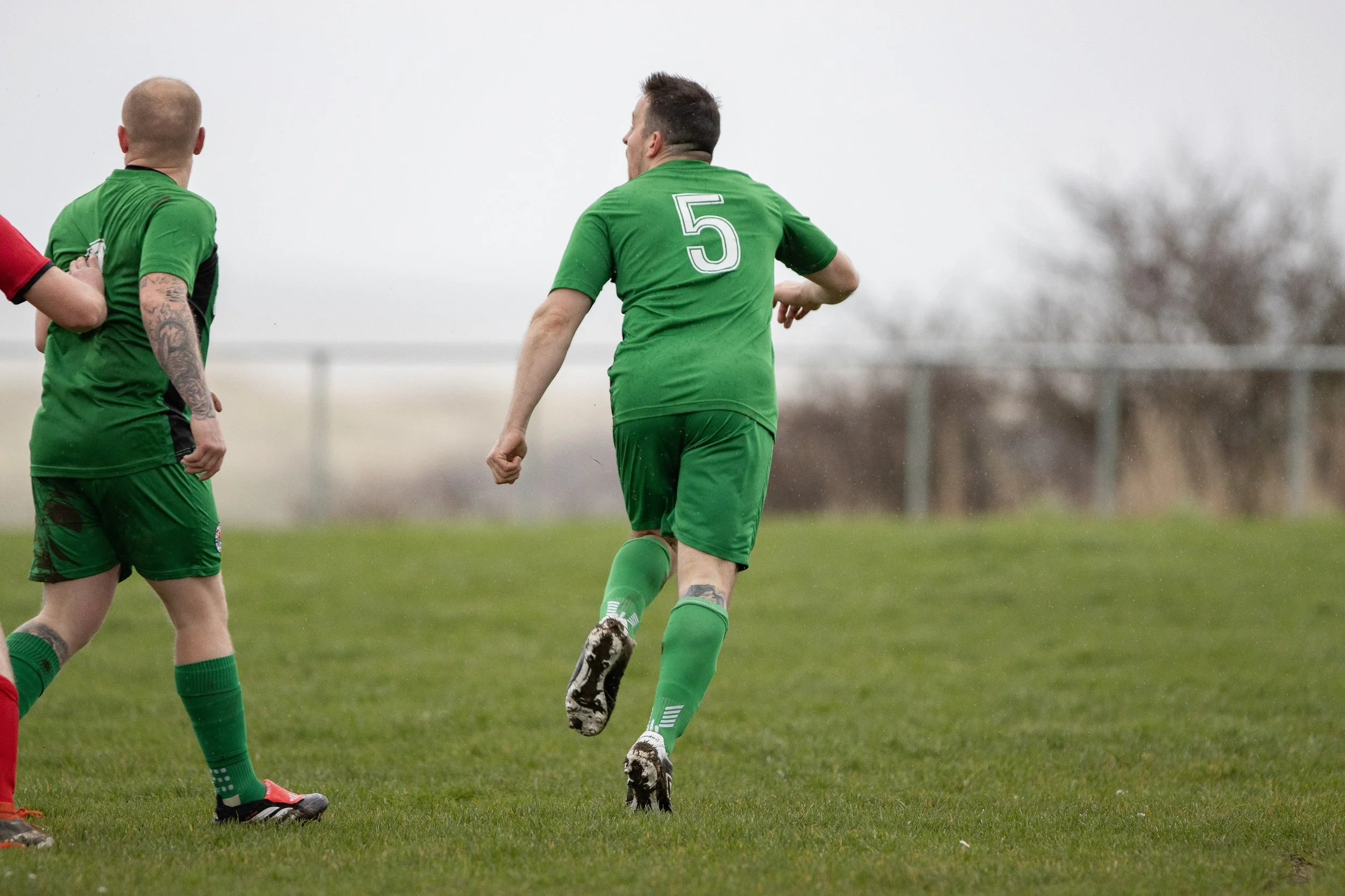 A soccer player in a green uniform with the number 5 on his back is running on a grassy field during a game on a cloudy day, with other players partly visible nearby.