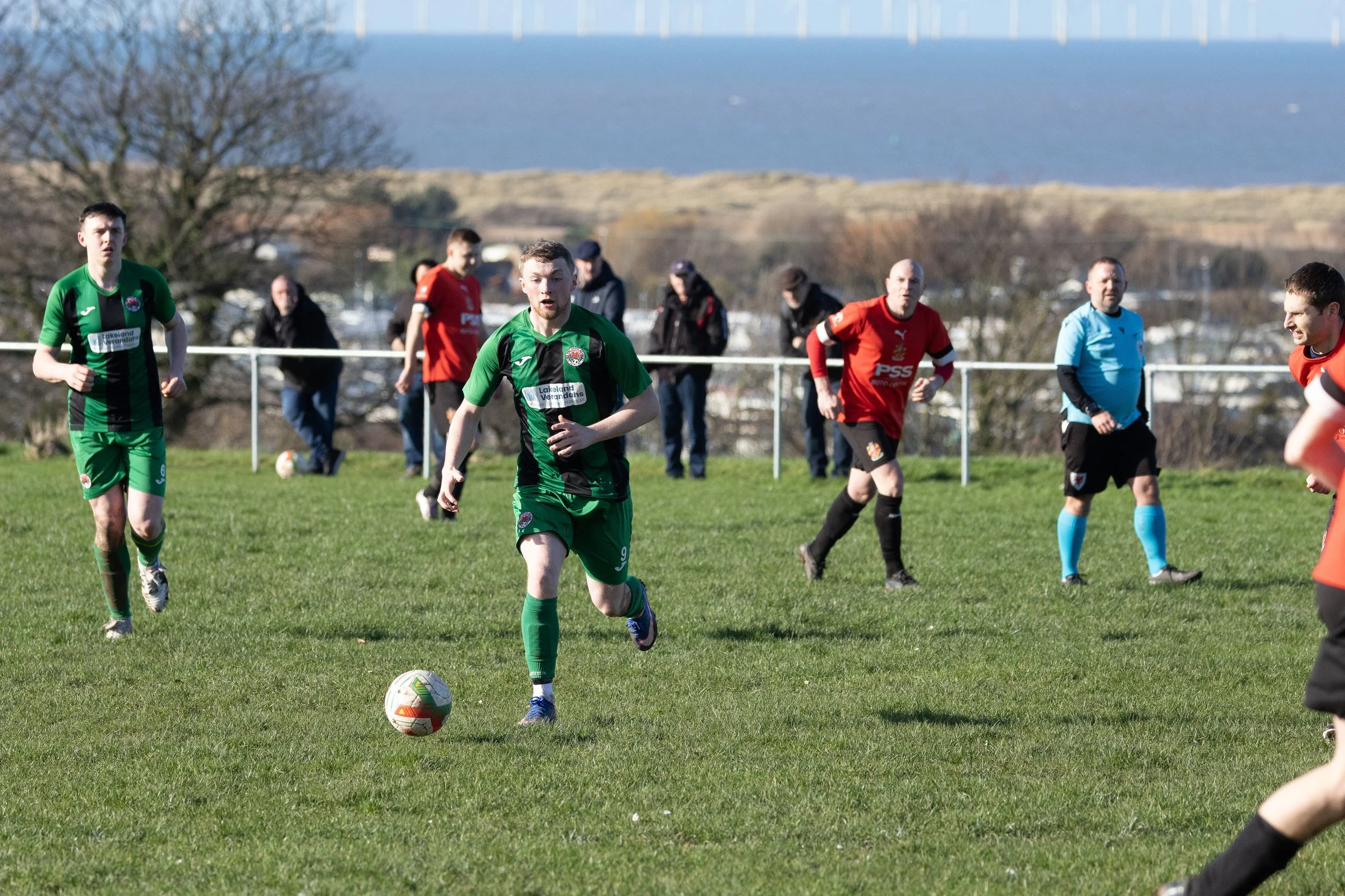Soccer match with players in green and red jerseys on a grassy field, with spectators in the background and a scenic landscape with water and hills behind.