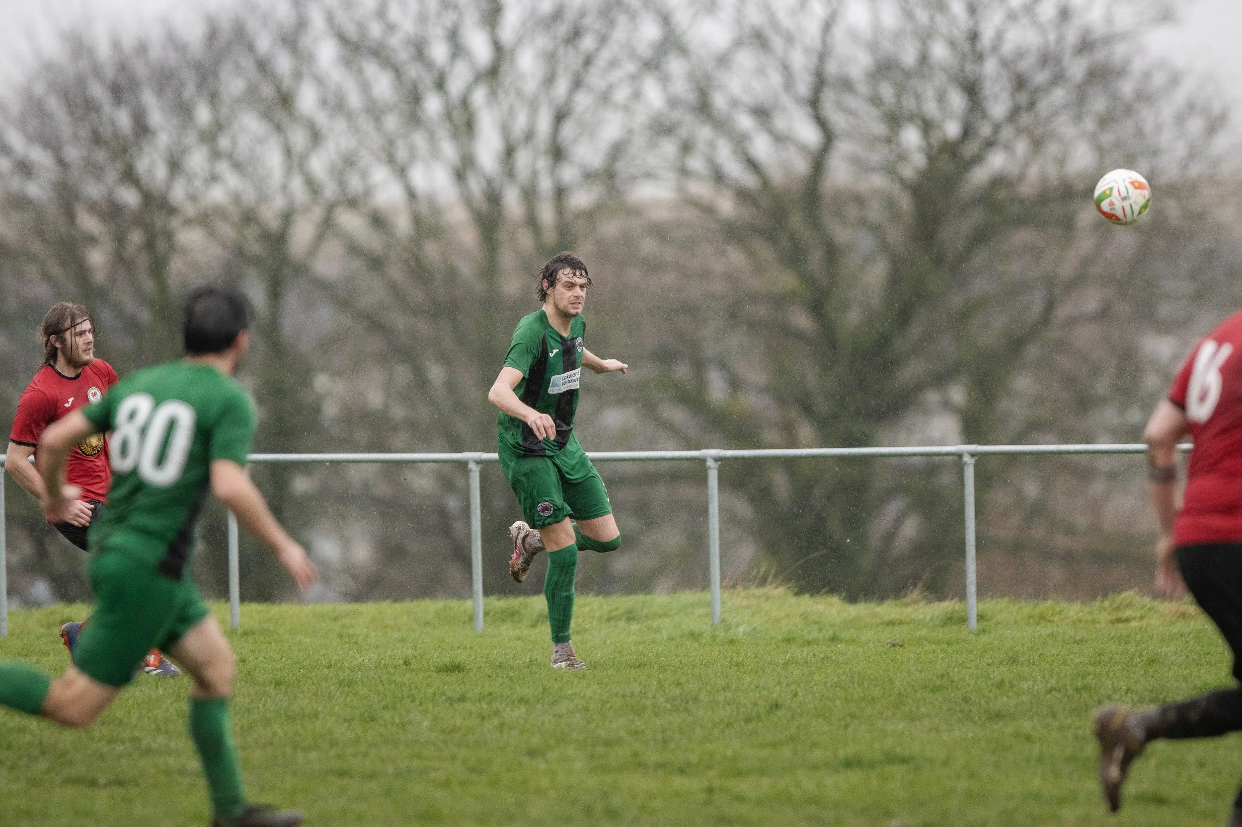Soccer players on the field during a game, with one in green about to head the ball, and others in red and green jerseys watching.