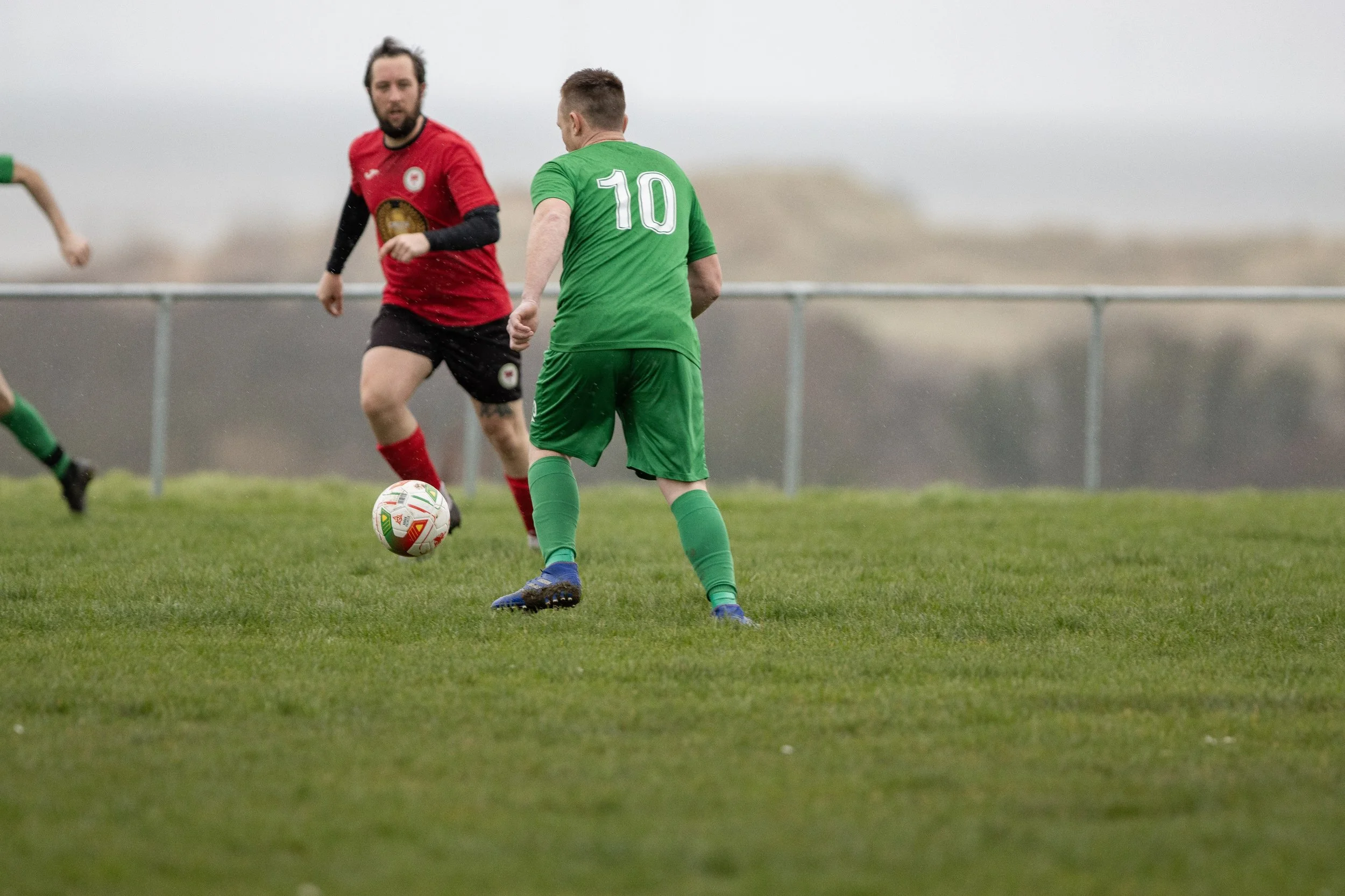 Two soccer players on a grassy field, one in a green uniform with the number 10, and the other in a red and black uniform, competing for the ball.