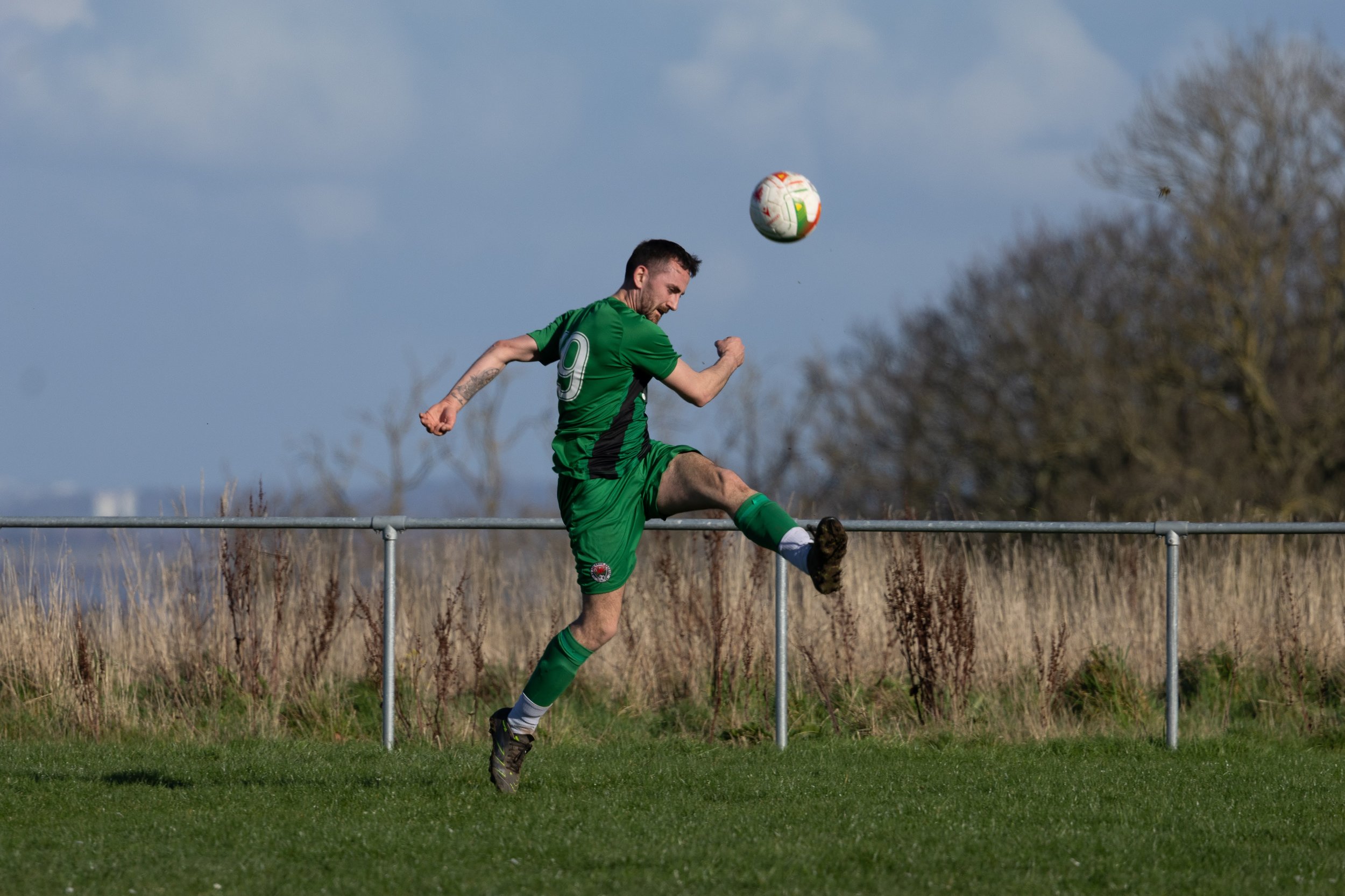 A soccer player in a green uniform kicking a soccer ball on a field with a metal fence and trees in the background.