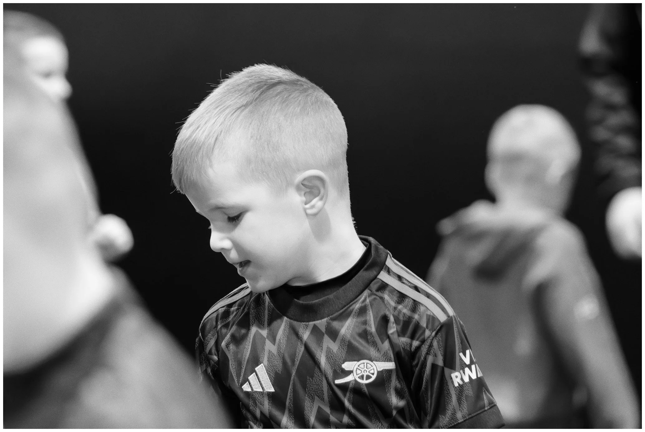 A young boy with short blond hair looking down, wearing a sports jersey with a logo of Arsenal football club.