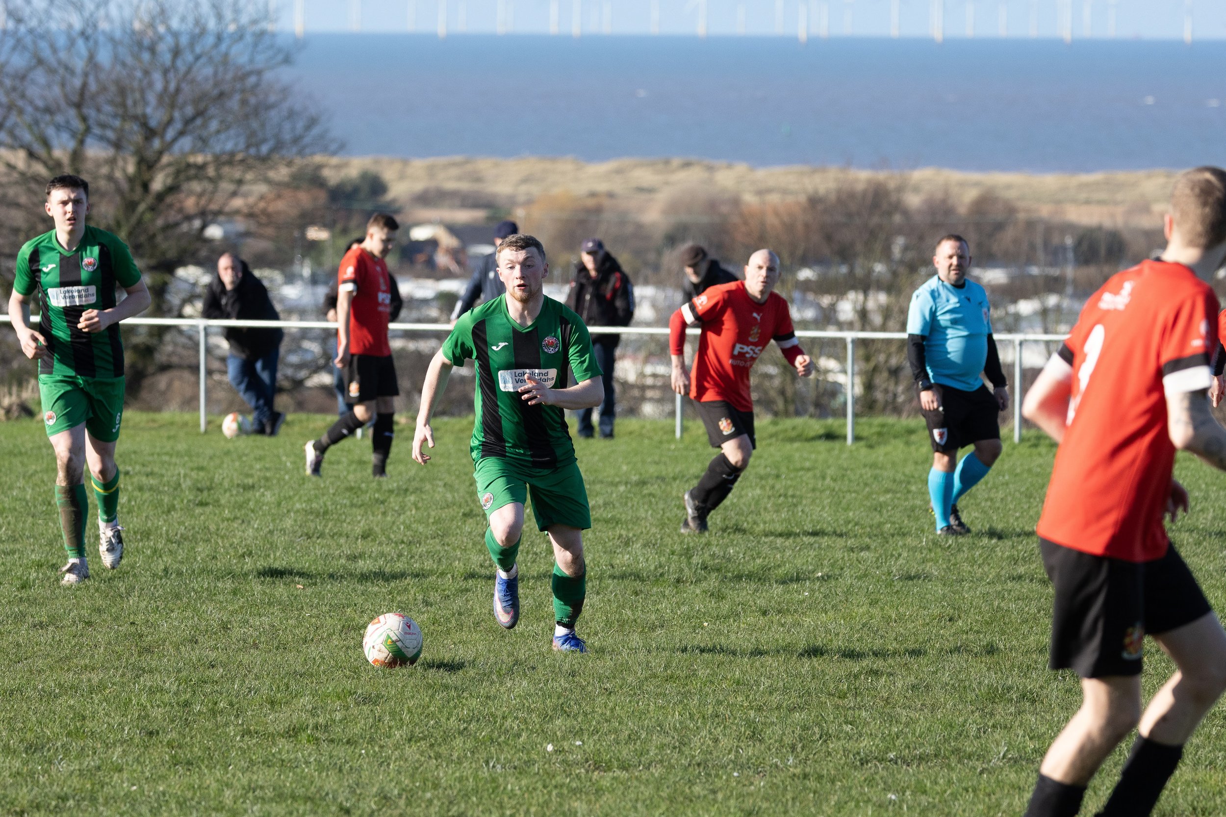 Soccer match with players in green and red jerseys on a grassy field, with a blue sky and hills in the background.
