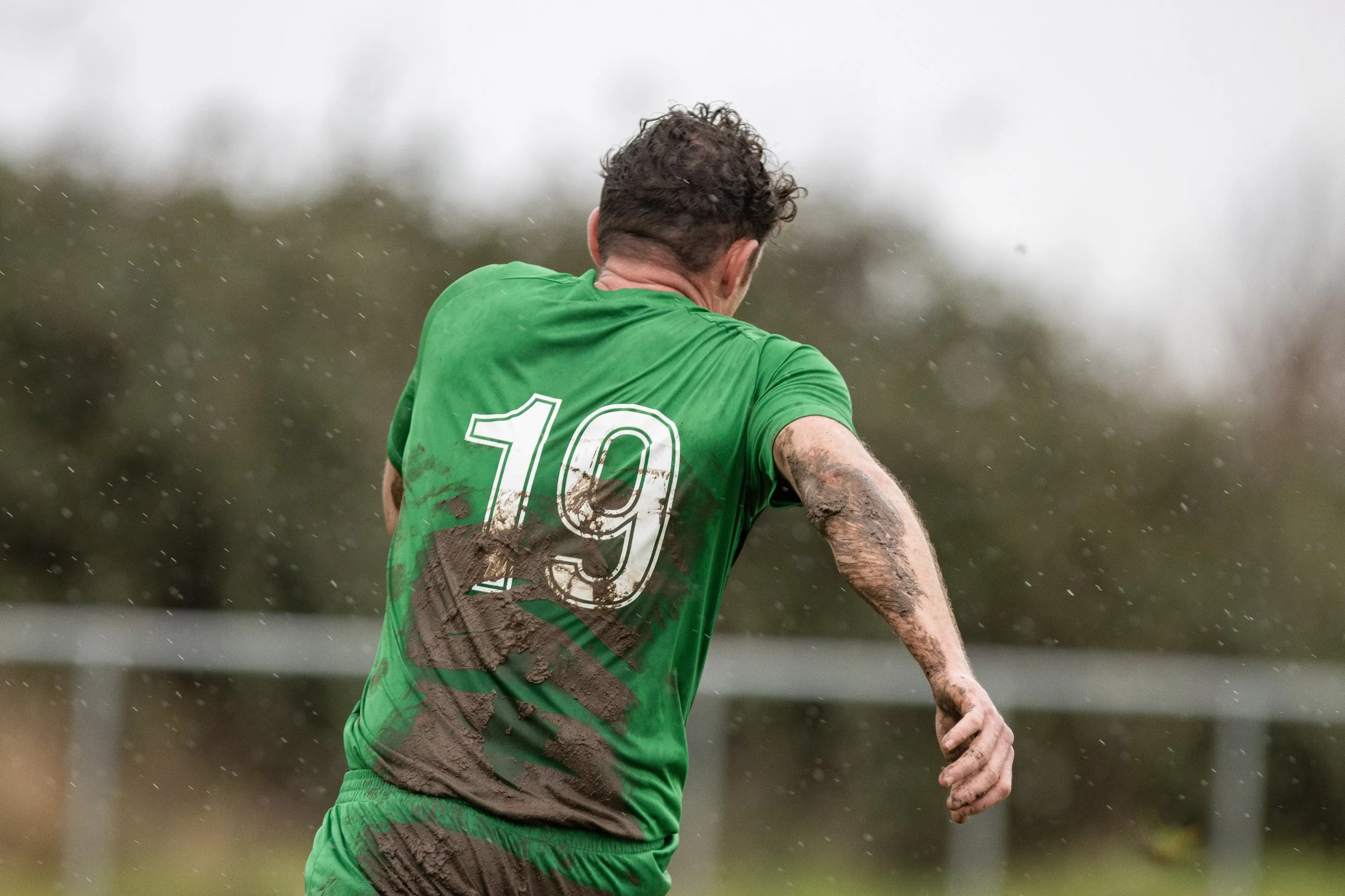 Back of a soccer player wearing a green jersey with the number 10, covered in mud, running on a muddy field in rainy weather.