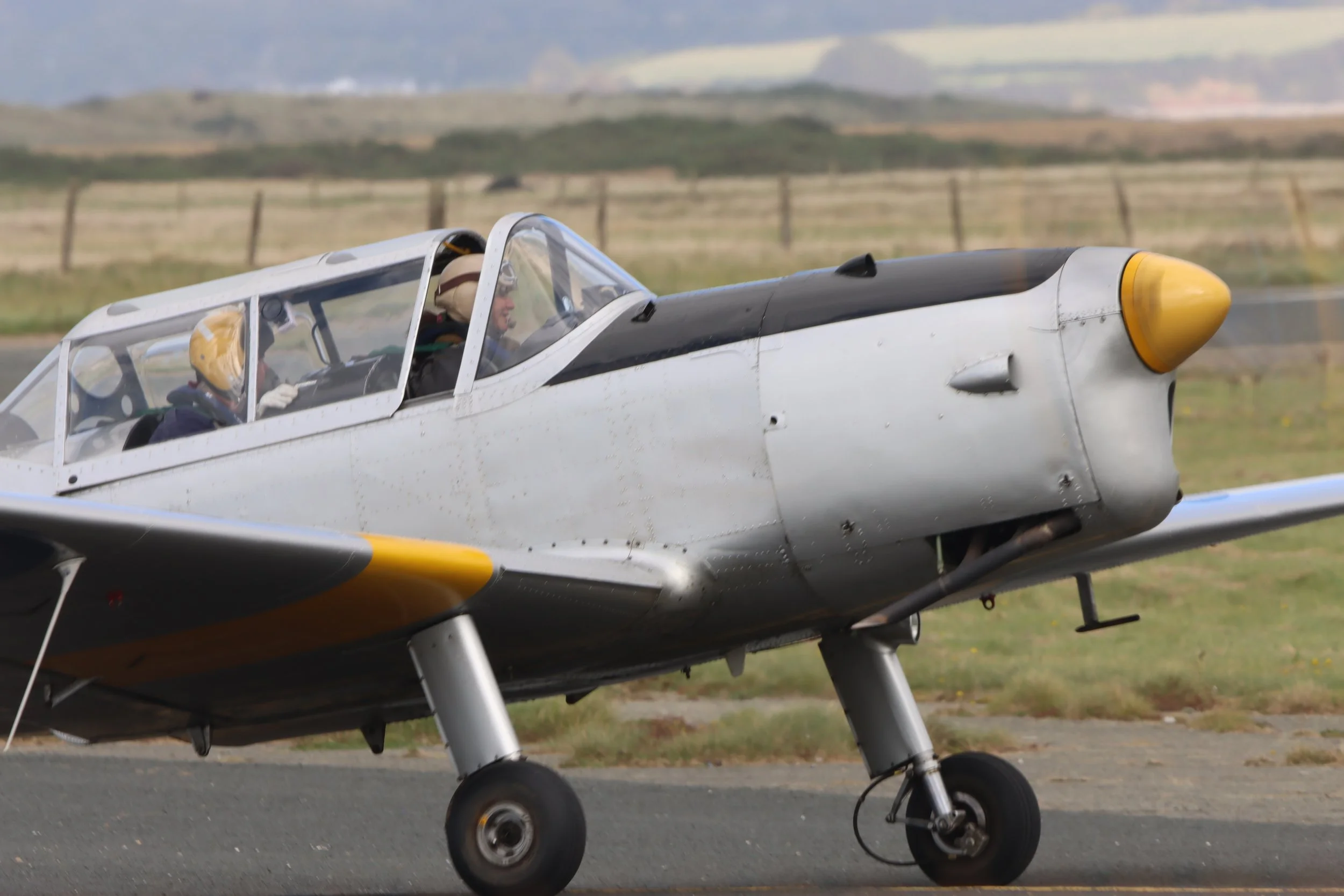 A vintage silver and black aircraft on a runway with two pilots wearing helmets in the cockpit, set against a rural landscape with rolling hills in the background.