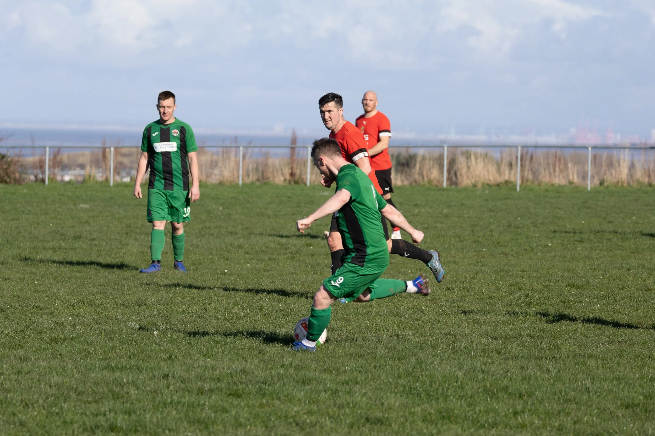 A soccer game with players in green and red uniforms on a grassy field, with players near the ball and a cloudy sky in the background