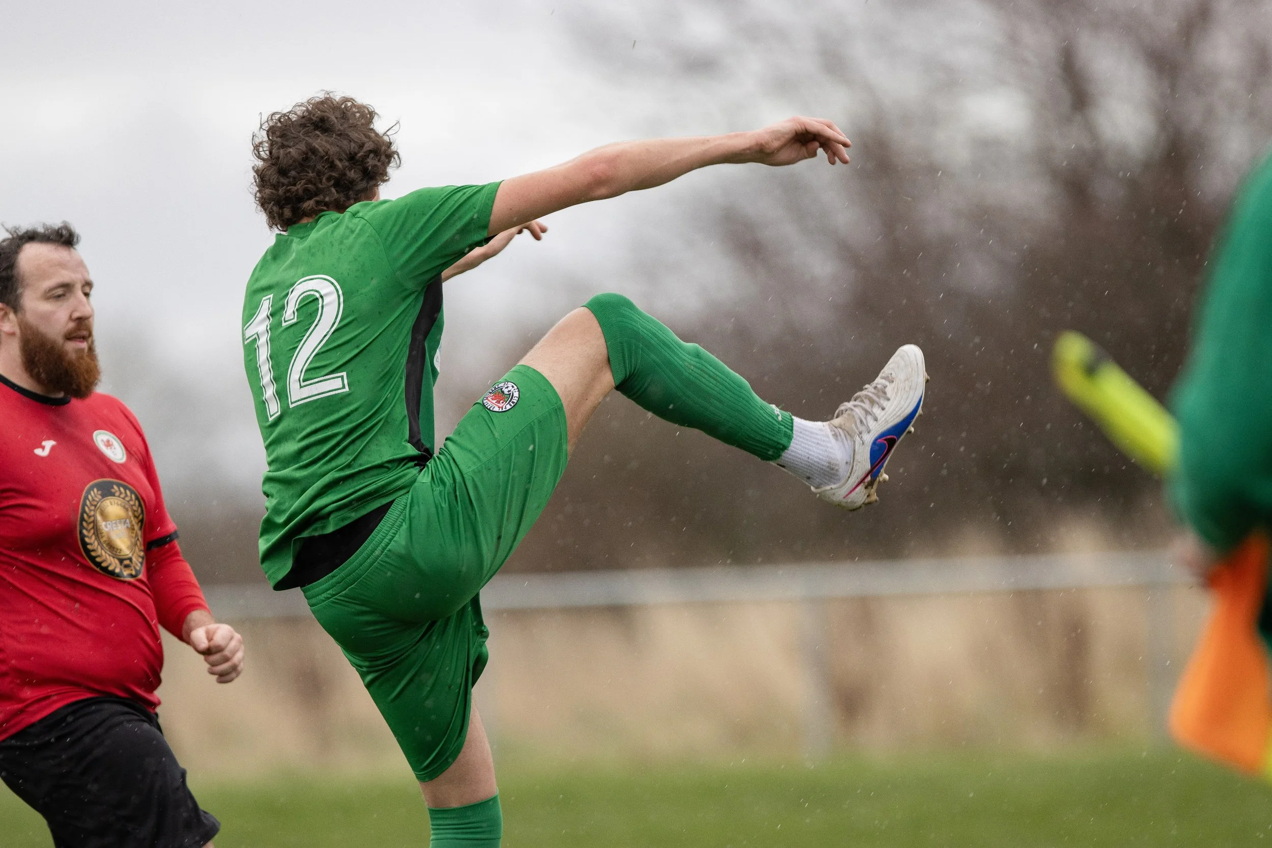 A soccer player in a green jersey with the number 12 kicking a ball during a match, with another player in a red jersey nearby on a rainy day.