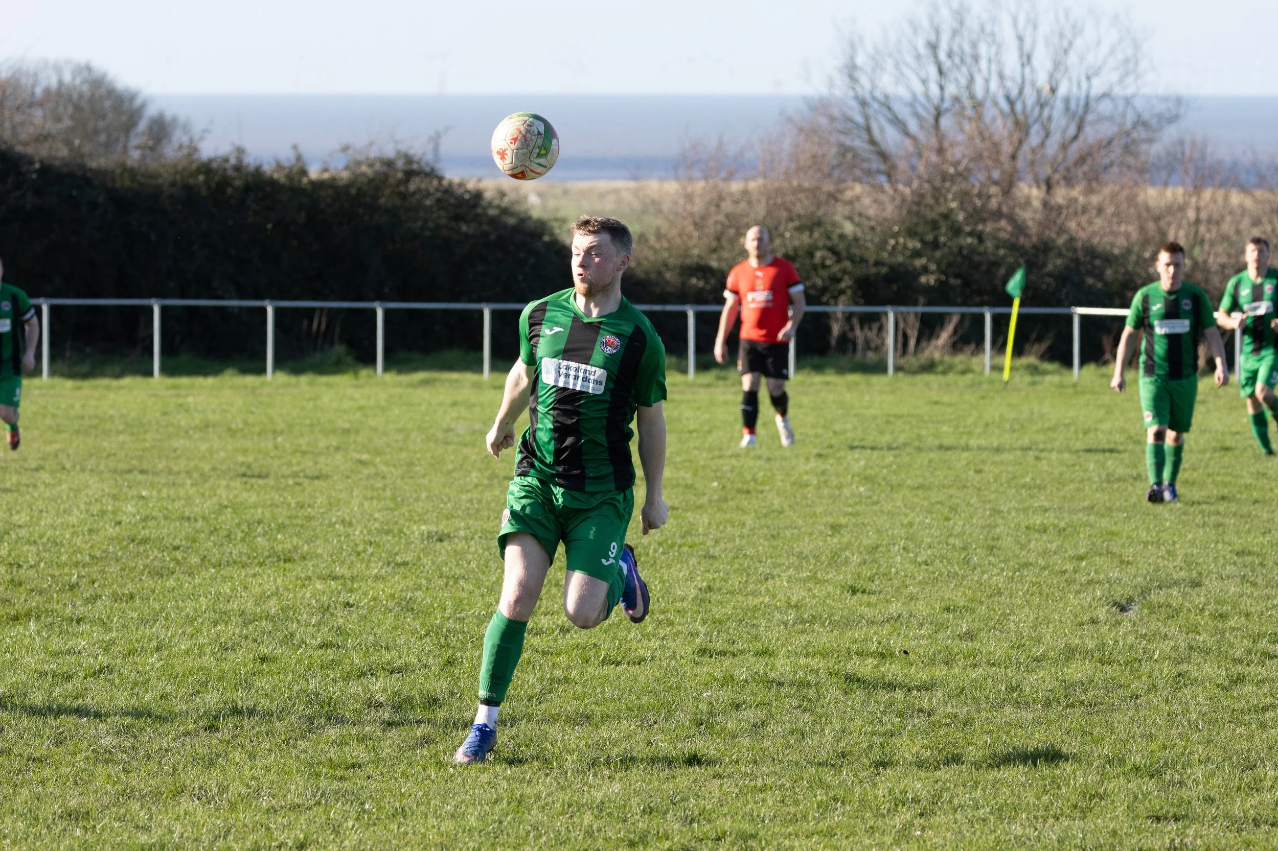 A soccer match with players in green and black uniforms on a grassy field, with a player in the foreground chasing the ball while others observe, and a background of trees and the ocean.