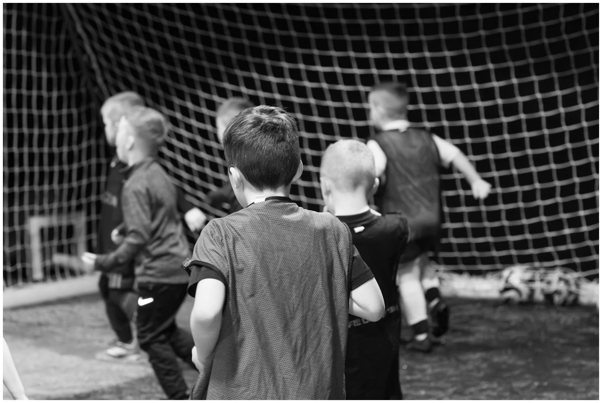 Young boys in sports jerseys standing in front of a soccer goal net, some facing away and some looking towards the net.