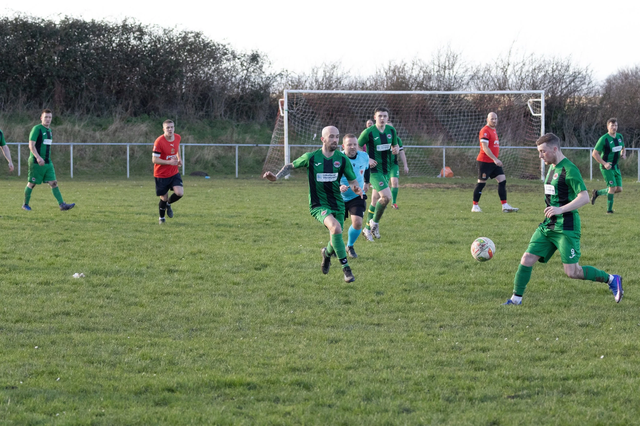 Soccer players on the field during a match, with some players running and others standing near the goalpost, on a grassy field with trees in the background.