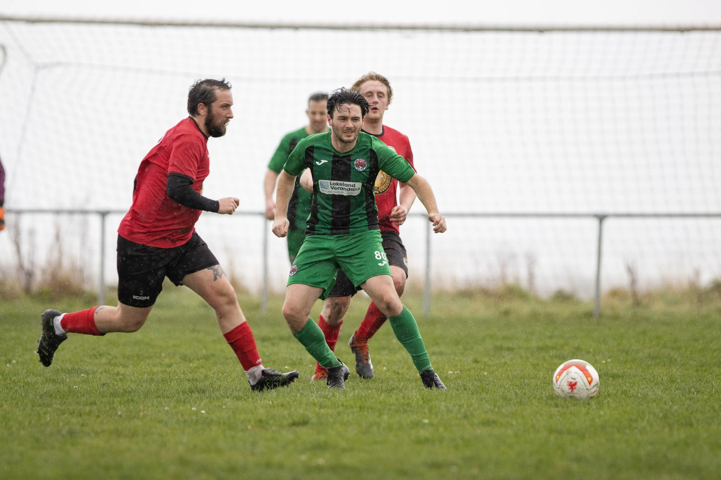 Group of soccer players in action during a match on a grassy field, with players in green and red uniforms chasing the ball, a soccer goal in the background.
