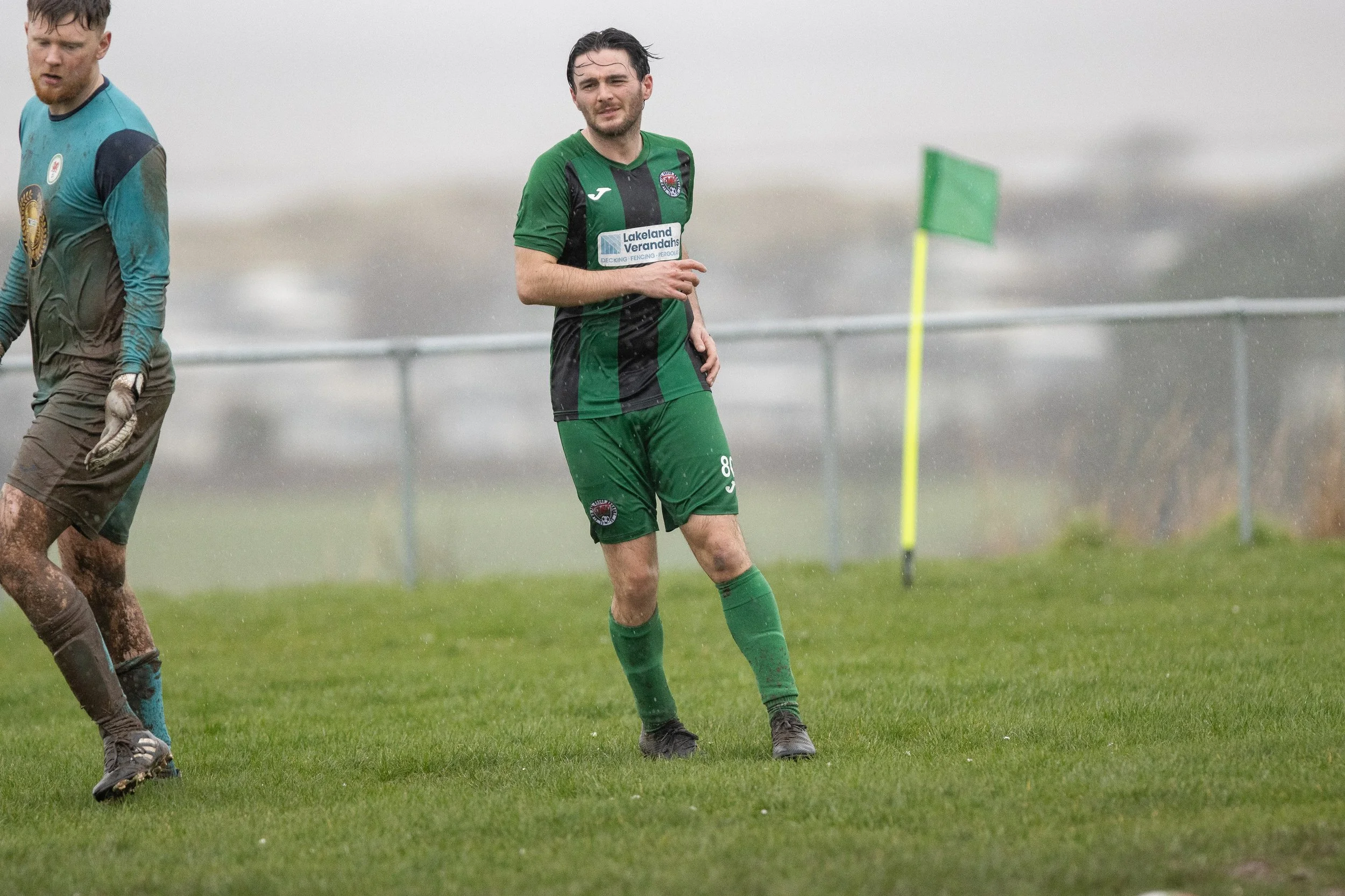 Soccer players on the field in rainy weather, one player in green uniform standing near a corner flag, and another player in blue and green goalkeeper attire to the left.