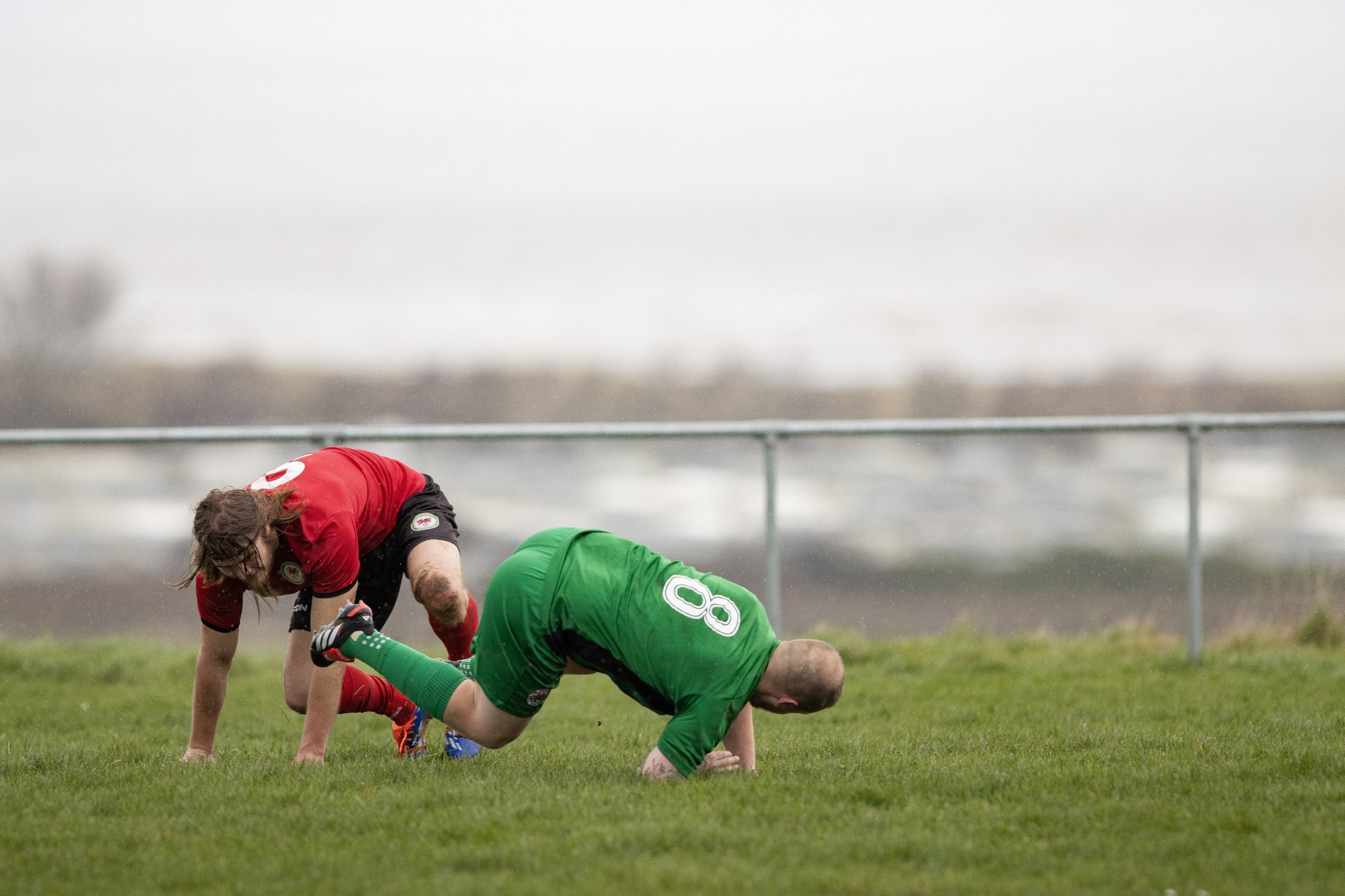 Two soccer players, one in a red jersey and one in a green jersey, are on the grass field, with the player in green appearing to be on all fours and the player in red bending over.