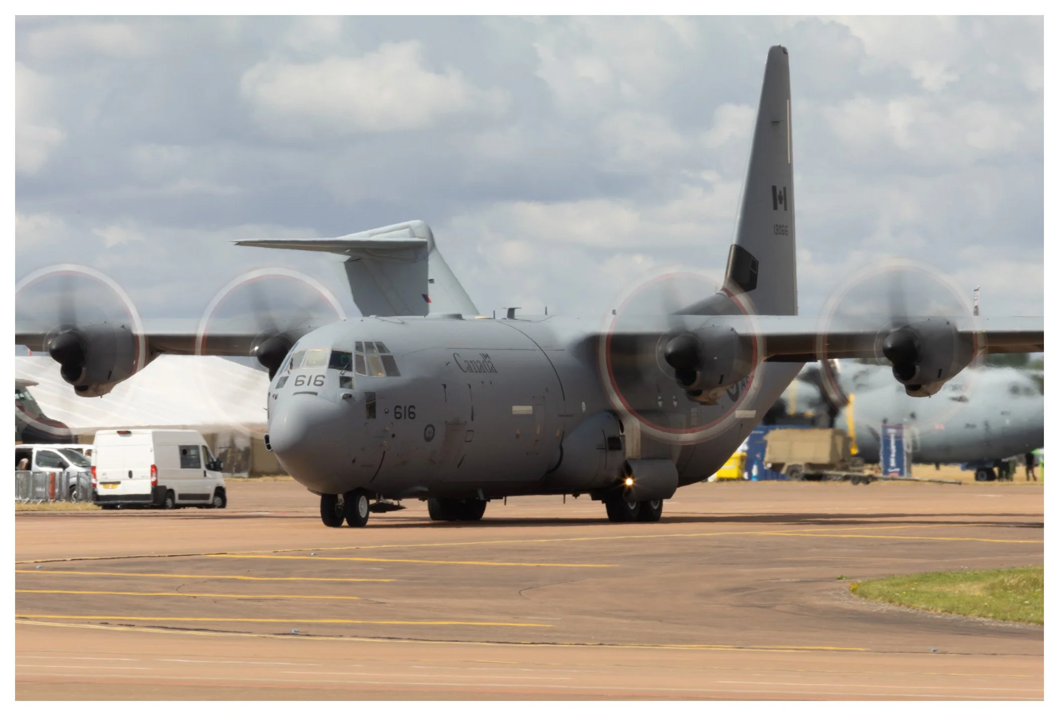 A military cargo plane on the runway with several propellers spinning, parked near other aircraft and vehicles.