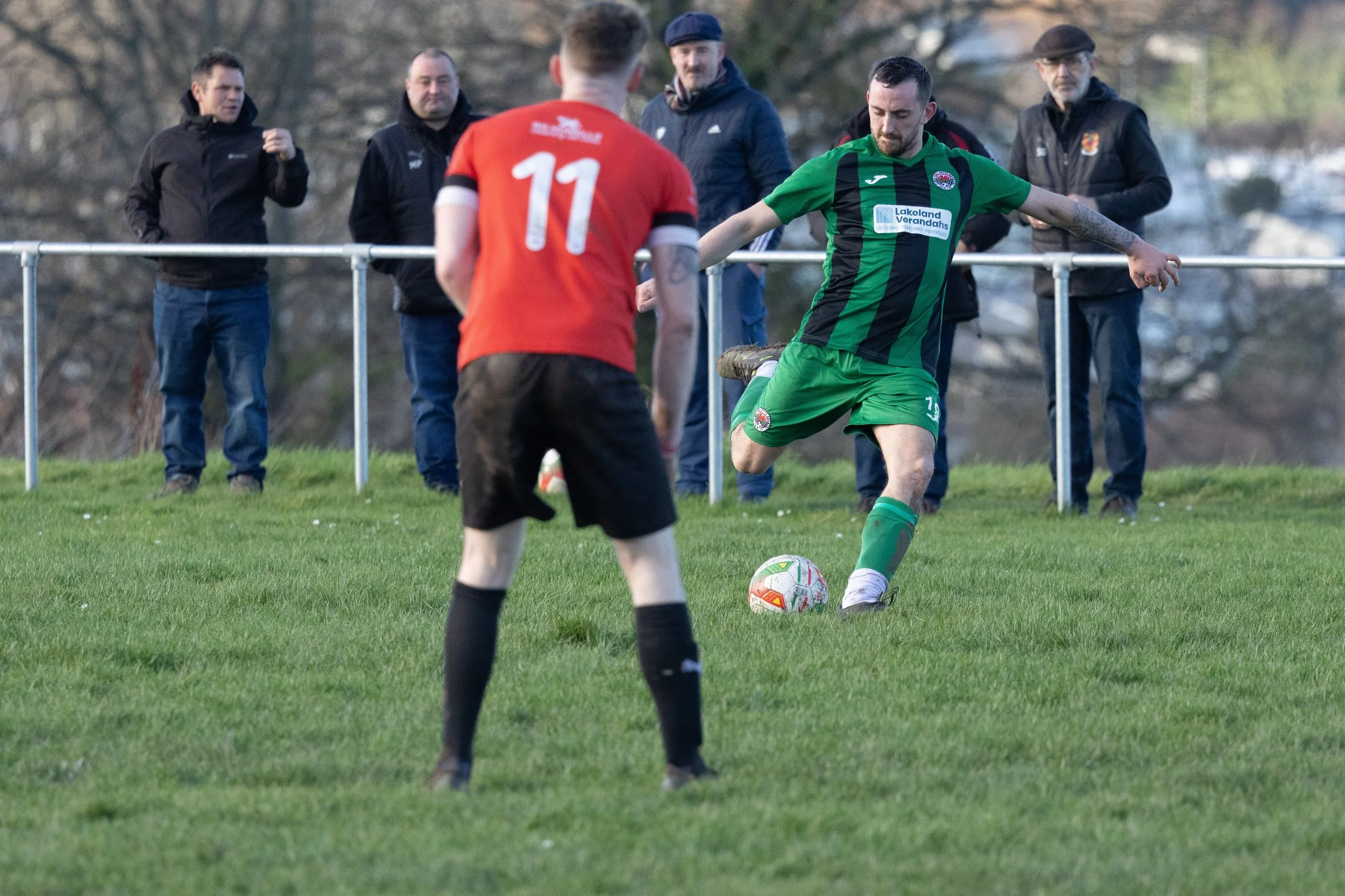A soccer player in a green and black kit is kicking a white soccer ball, with a player in a red jersey numbered 11 standing nearby on a grassy field, and four spectators watching behind a metal fence in the background.