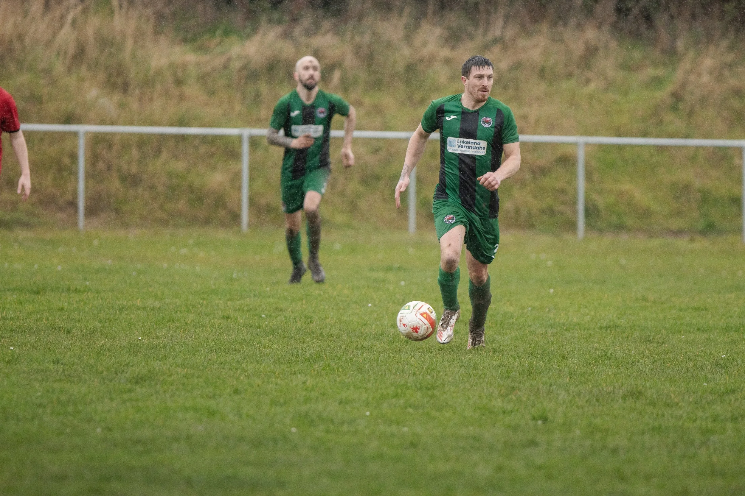 Soccer players on a wet field during a game, with one player in green kicking a ball and others running behind.