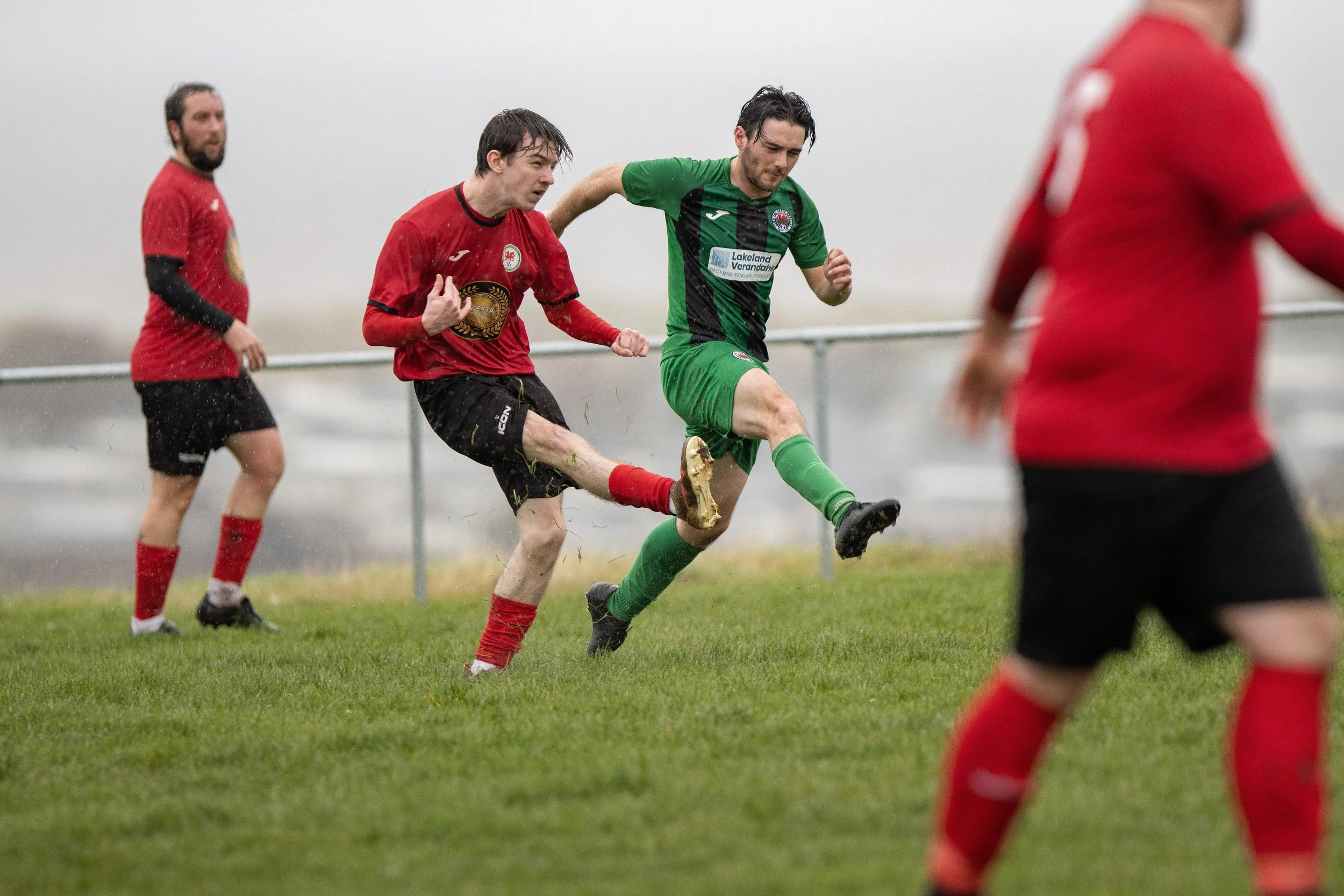 Soccer players in red and green uniforms competing for the ball on a rainy field.