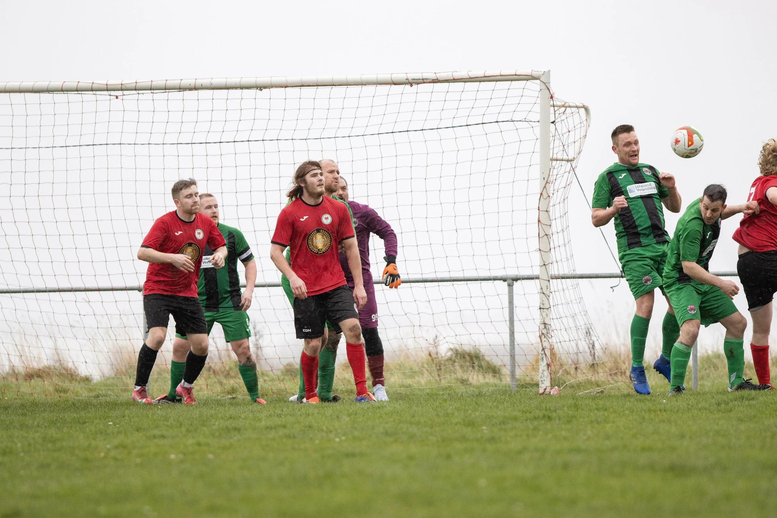 Soccer players in red and green jerseys competing near the goal during a game on a grassy field under an overcast sky.