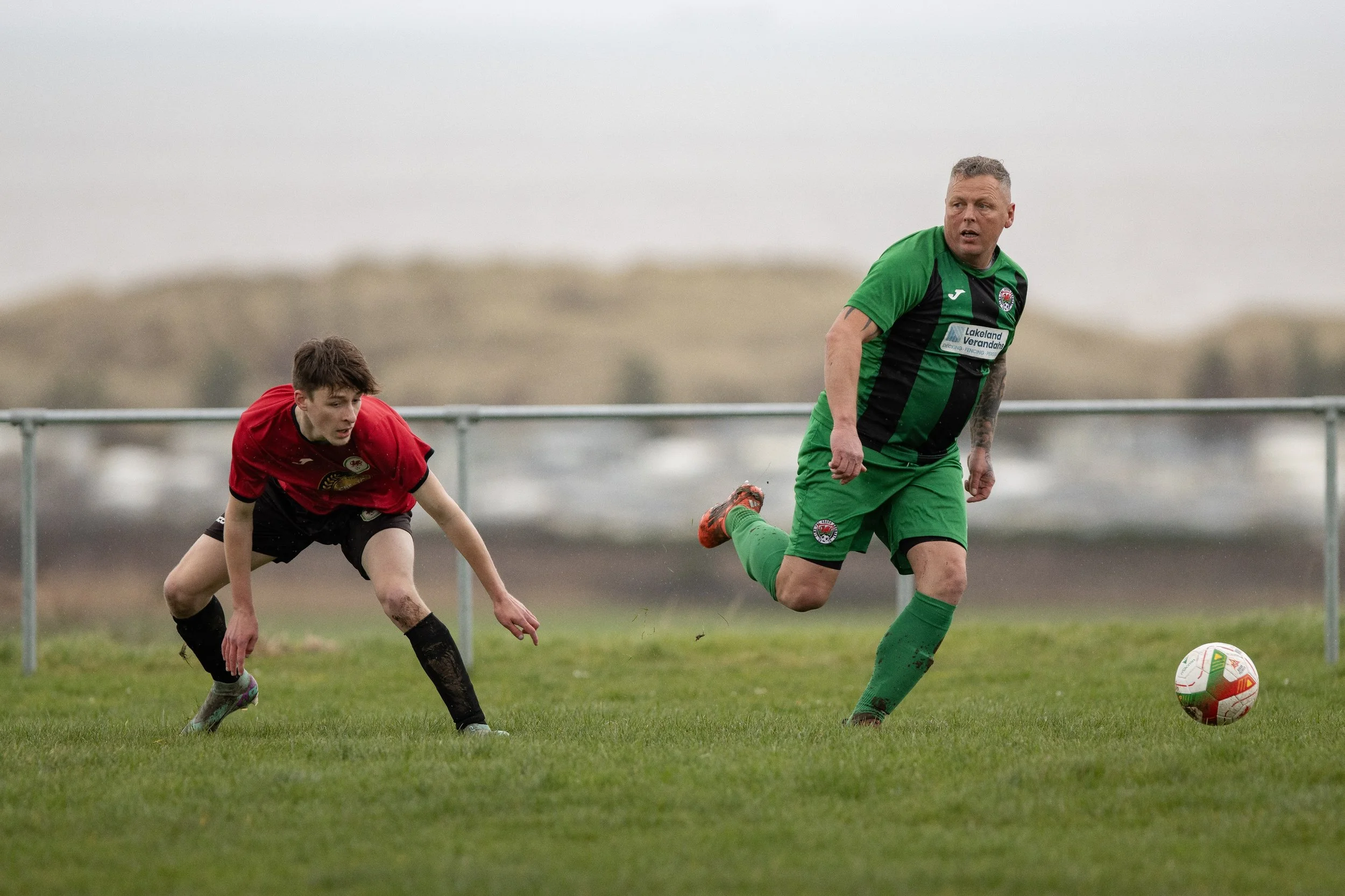 Two soccer players in action; one in a red jersey falling forward on grass field, the other in a green and black jersey running after a soccer ball on a cloudy day.