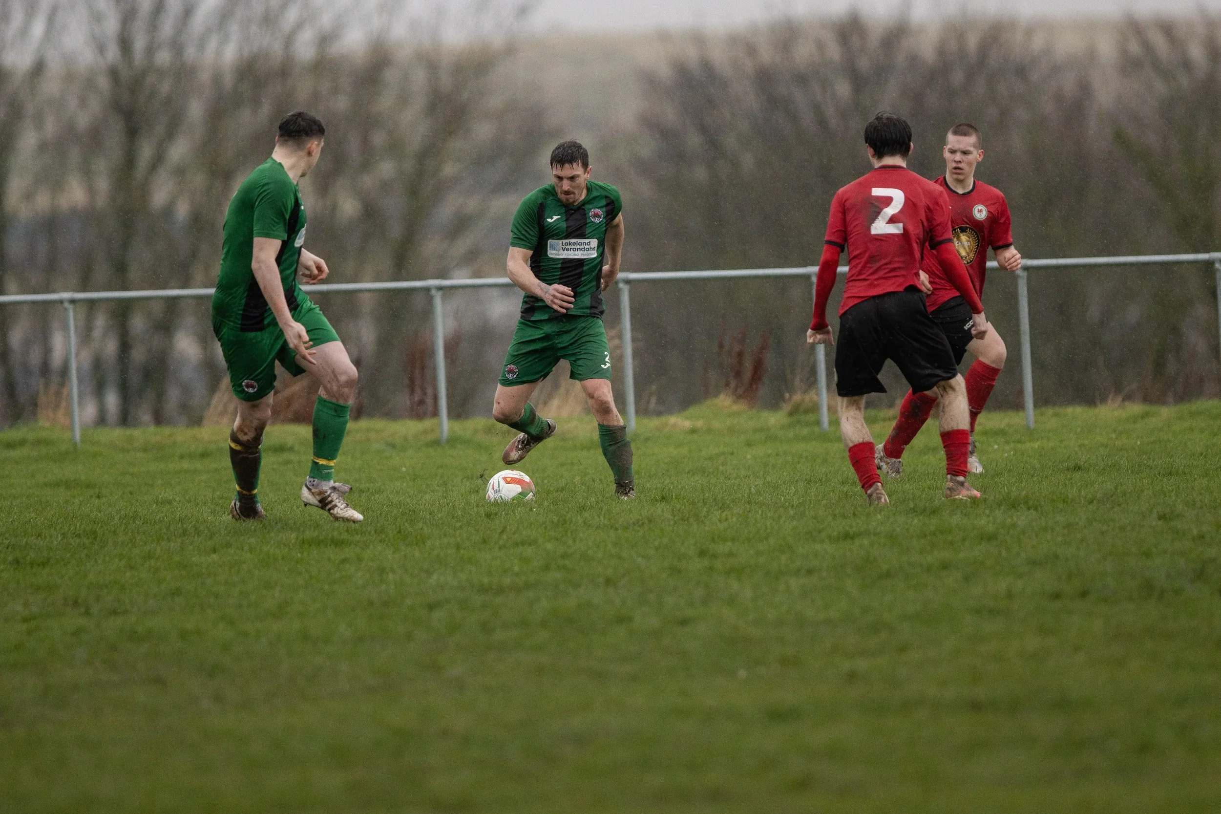 Four soccer players on a grassy field during a match, with trees in the background. One player in a green uniform is preparing to kick the ball, while two players in red uniforms are positioned nearby and another in green is observing.
