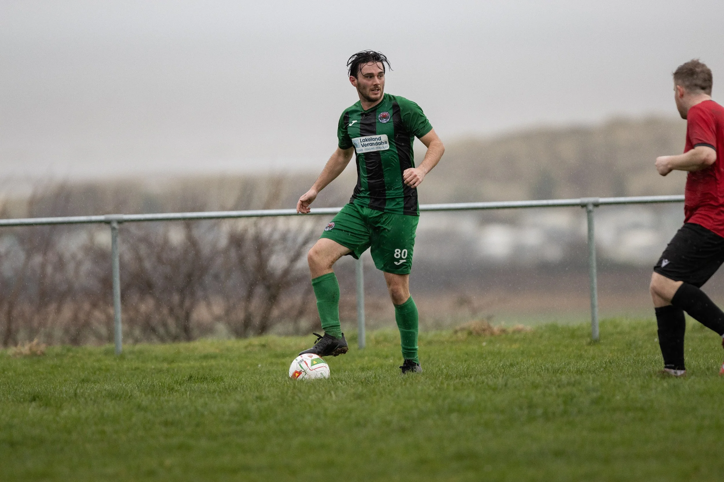 A soccer player in a green and black jersey with the number 80 controls the ball on a wet grassy field during a game, with another player in a red shirt approaching.
