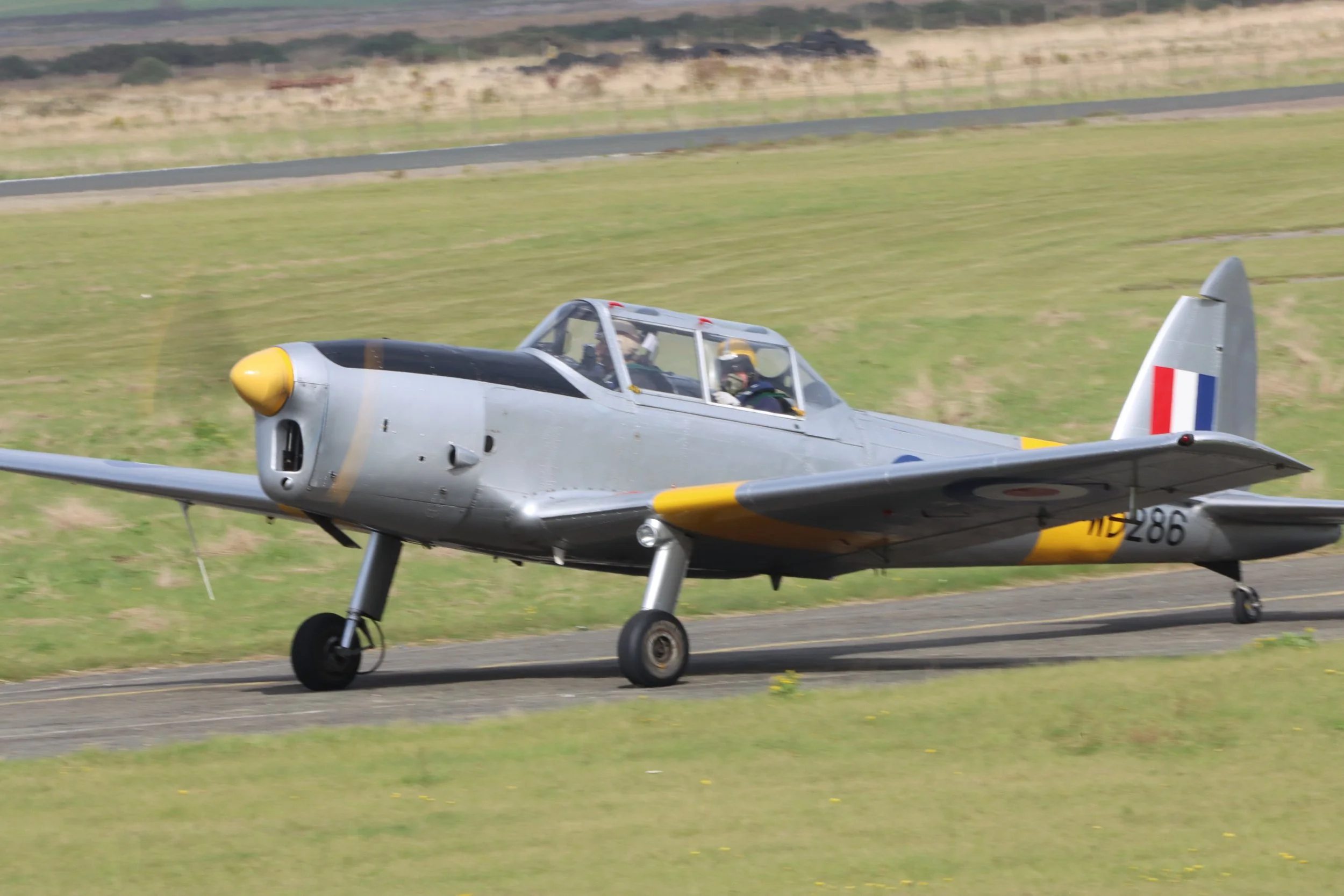 A vintage gray military aircraft with a yellow nose and a red, white, and blue roundel on the side, moving on a runway with green grass and a distant landscape in the background.