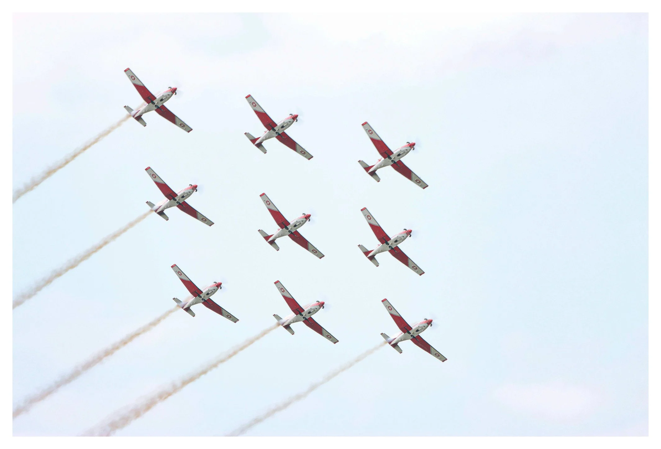 A formation of nine small red and white jet airplanes flying in a delta formation in the sky, leaving white smoke trails.