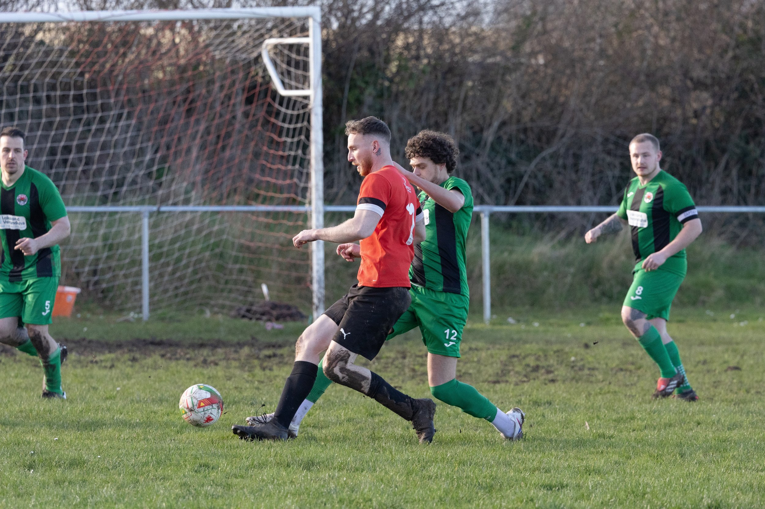 Soccer players on a grass field in action during a match, with a goalpost and net in the background. One player in a red jersey is competing with a player in a green jersey for the ball, while two other players in green are nearby.