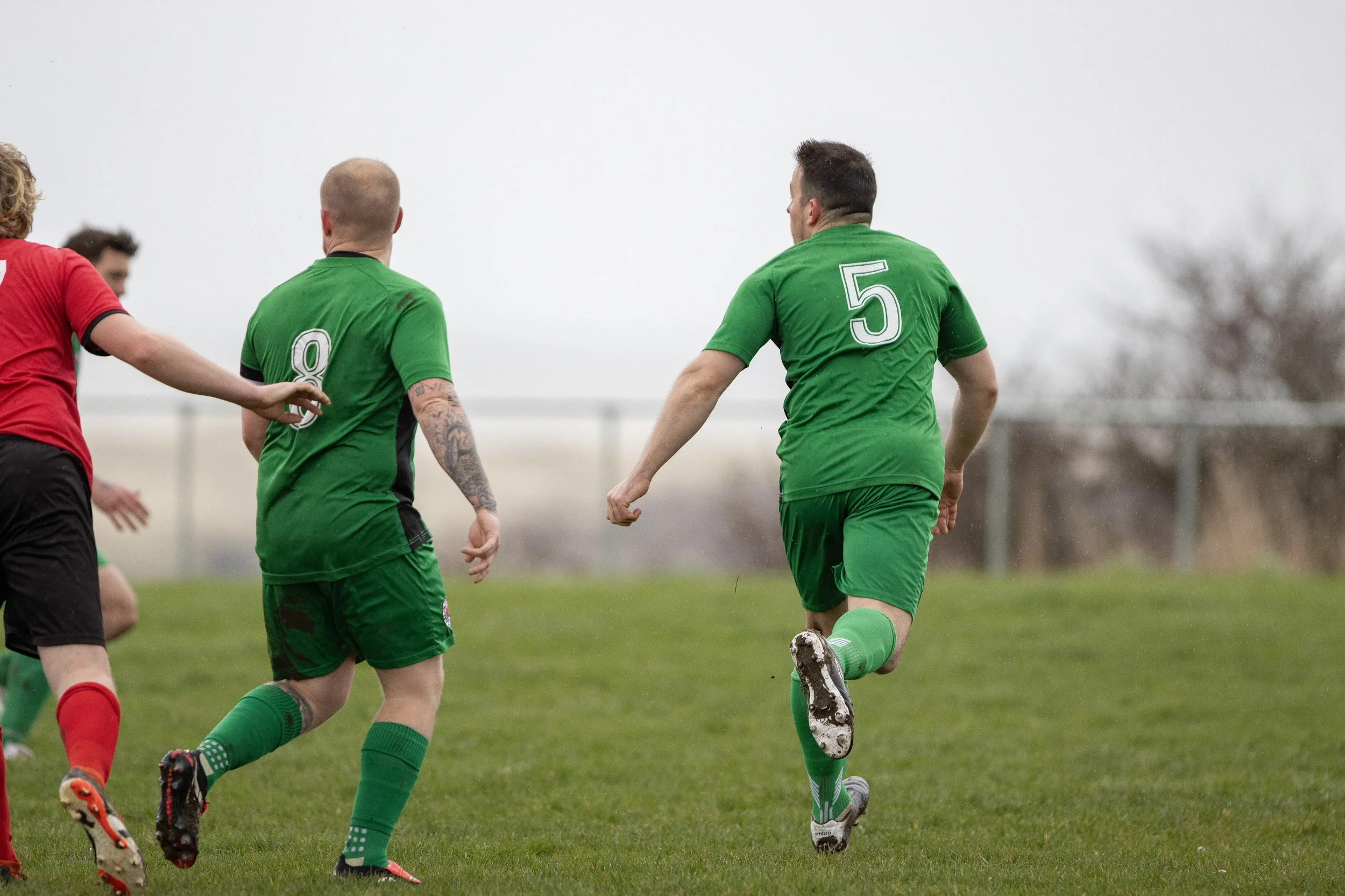 Soccer players in green and red jerseys during a match on a grassy field under an overcast sky.