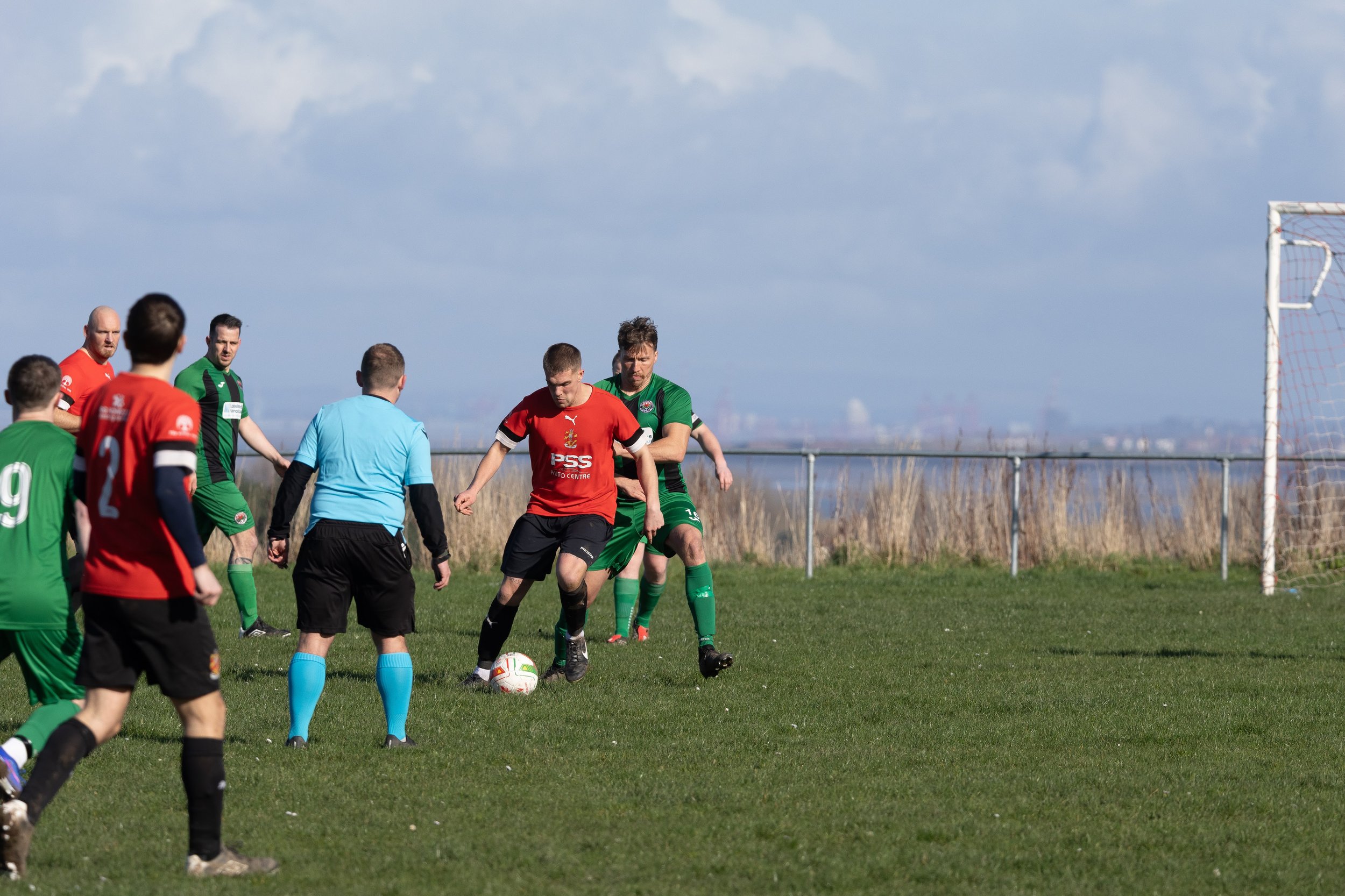 Soccer game with players and referee on a grassy field, players in red and green jerseys near the goal, with a fence and blue sky in the background.