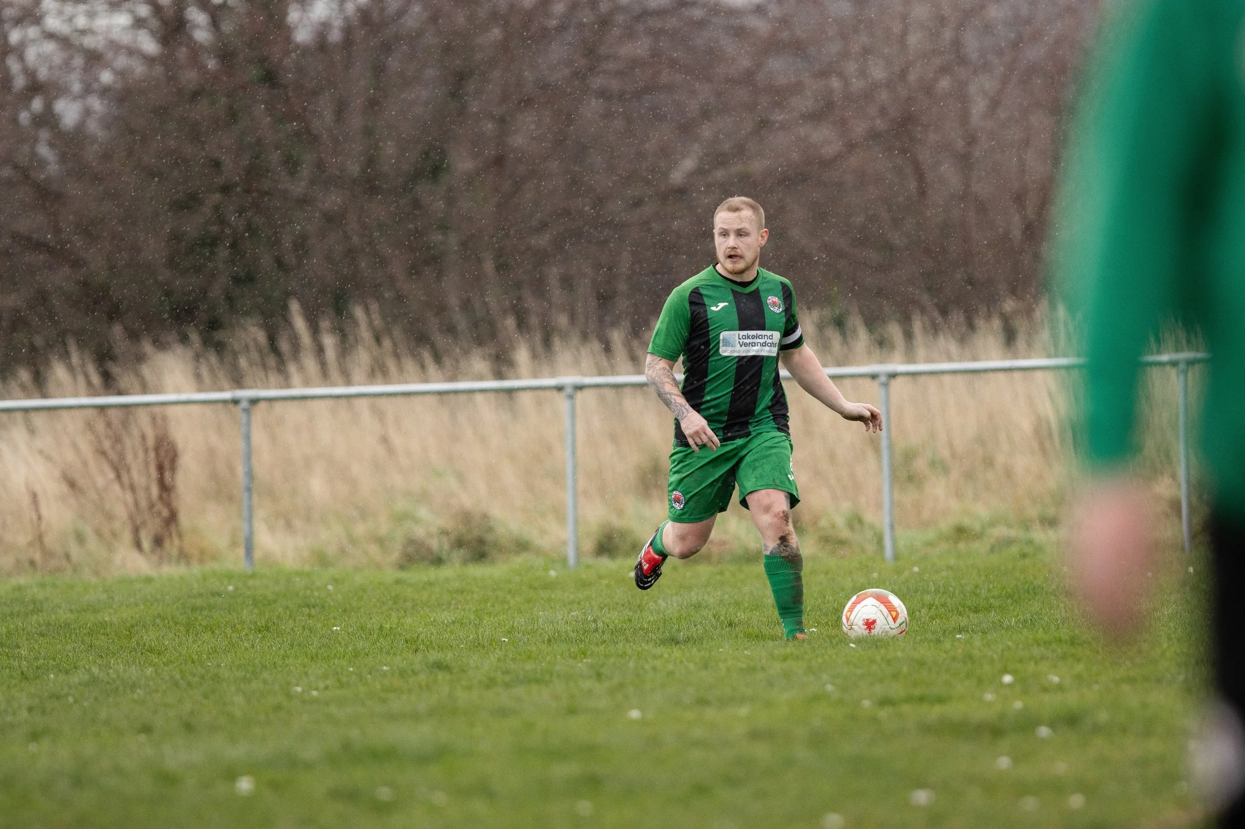 A male soccer player in a green and black uniform running on a grassy field with a soccer ball at his feet, during a match on a cloudy day.