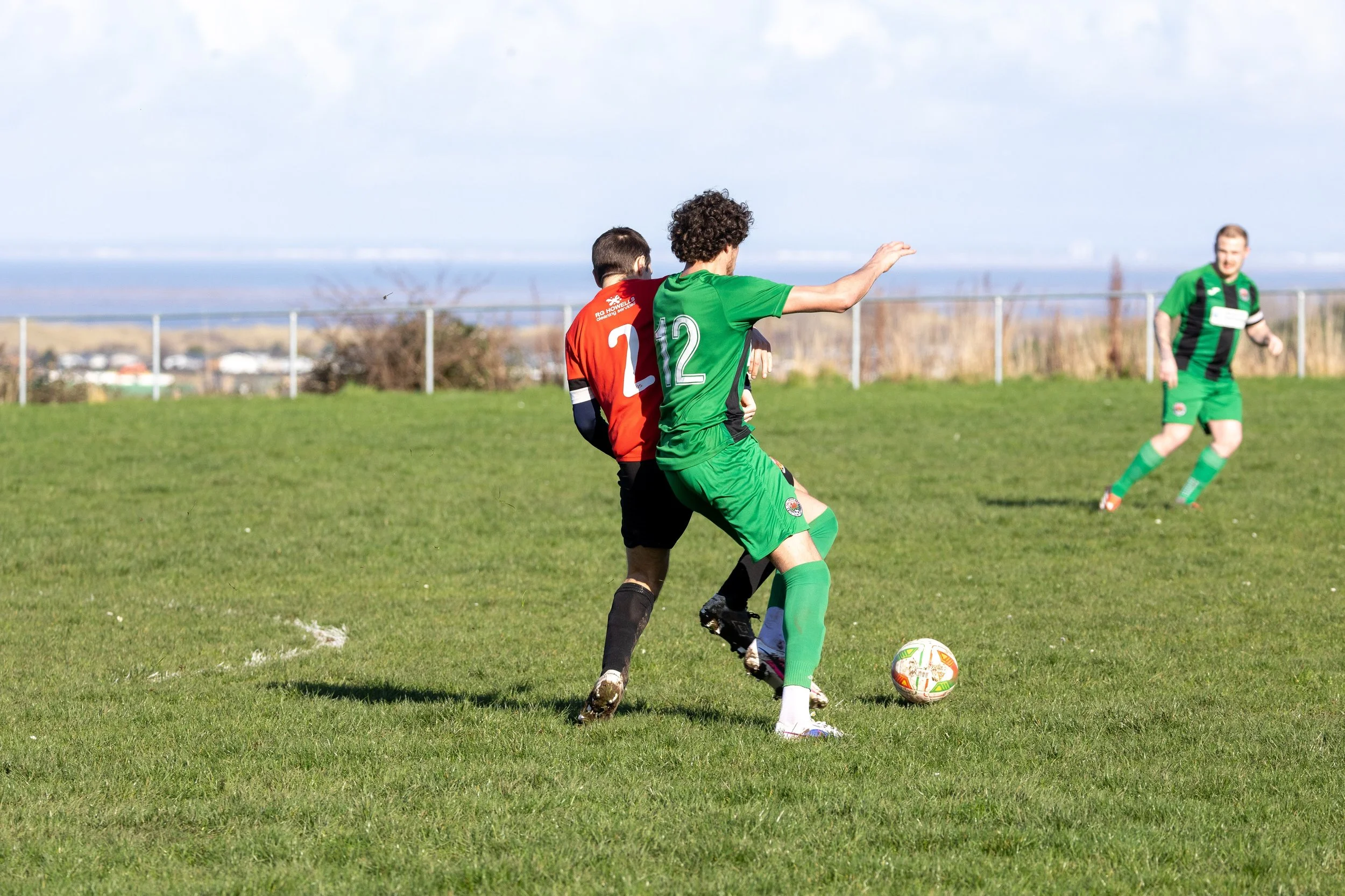 Two soccer players, one in red and black and the other in green, compete for the ball on a grassy field during a match, with another player in green observing in the background.