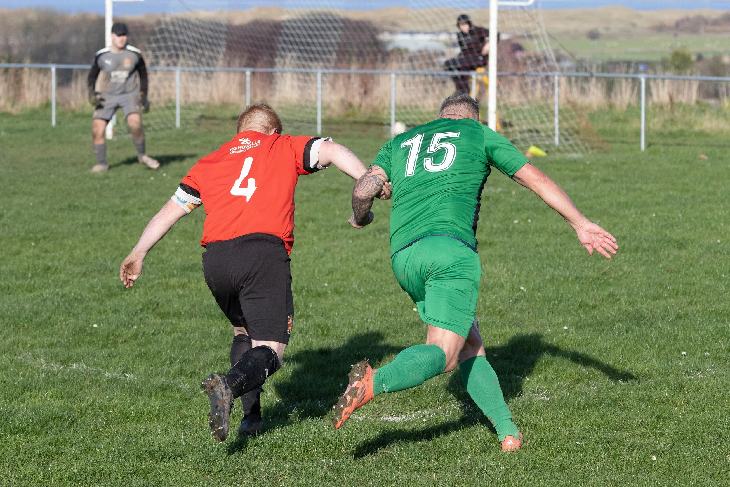Two soccer players, one in a red jersey with the number 4 and the other in a green jersey with the number 15, are running on a grassy field during a match. A goalkeeper is visible in the background near the goal, and a person sits on the goal post. T