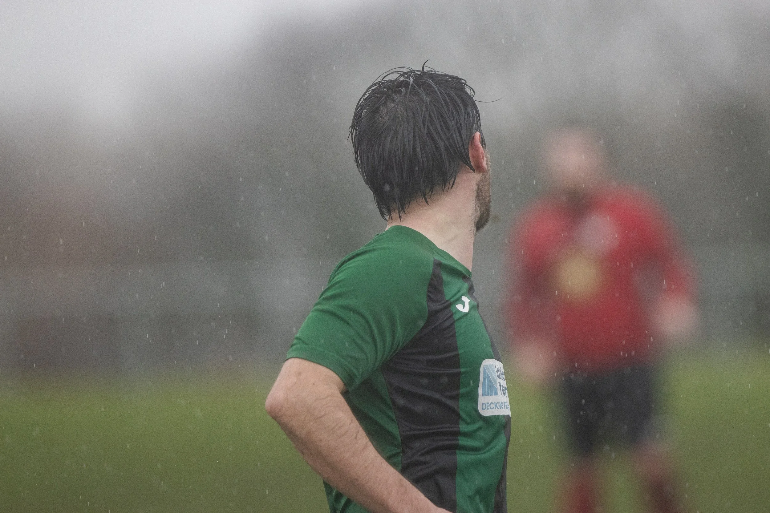A soccer player with black hair, wearing a green and black jersey, standing in the rain with his back to the camera, and another player in a red shirt blurred in the background.