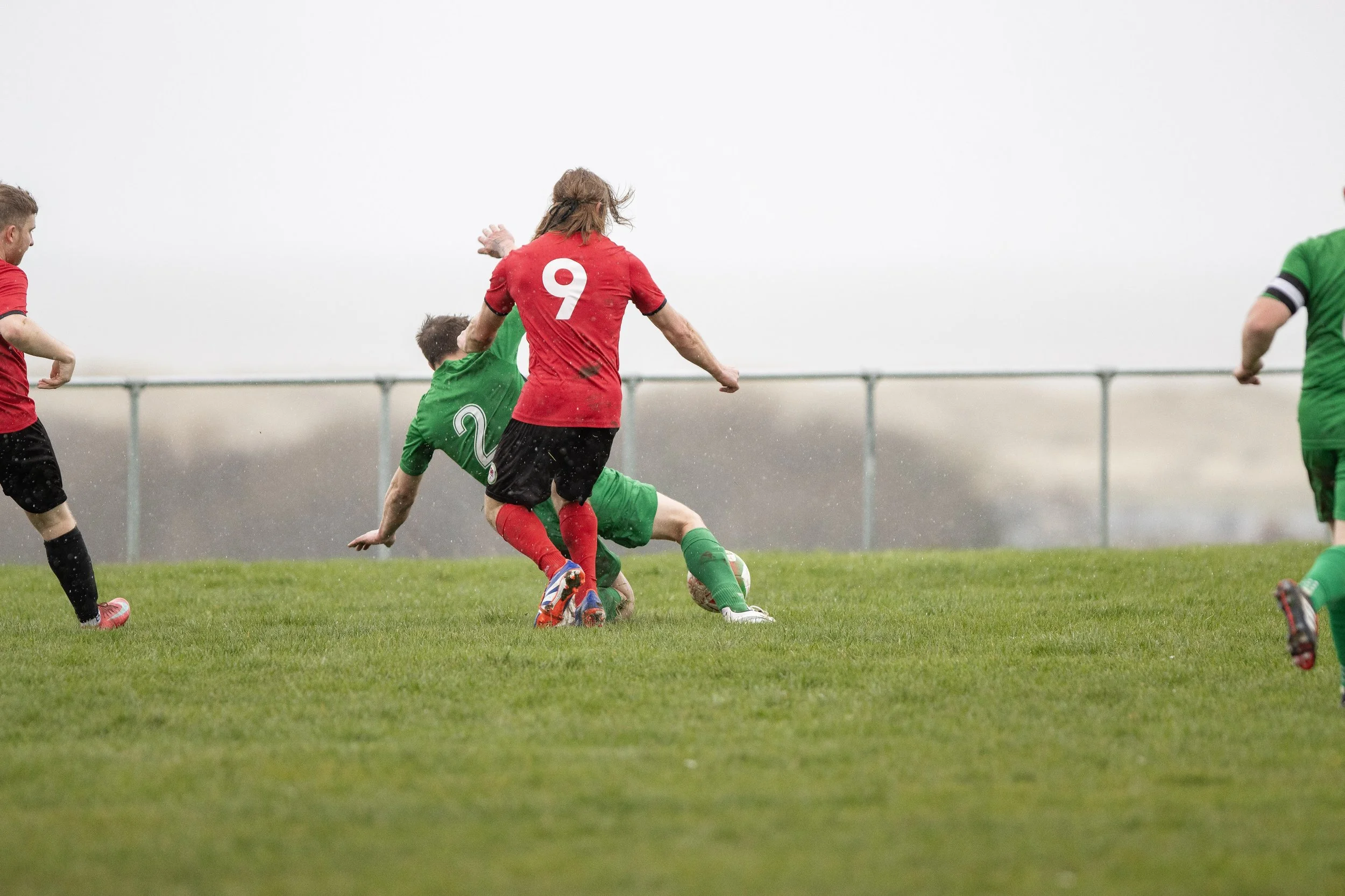 Soccer players in green and red jerseys competing for the ball on a grassy field in rainy weather.