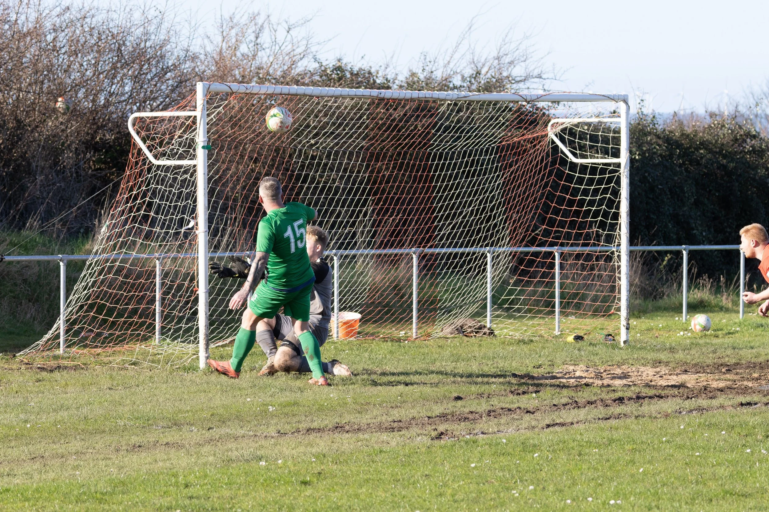 Soccer players contest a ball near the goal, with one in a green jersey and a goalkeeper in black and gray, on a grassy field during daytime.
