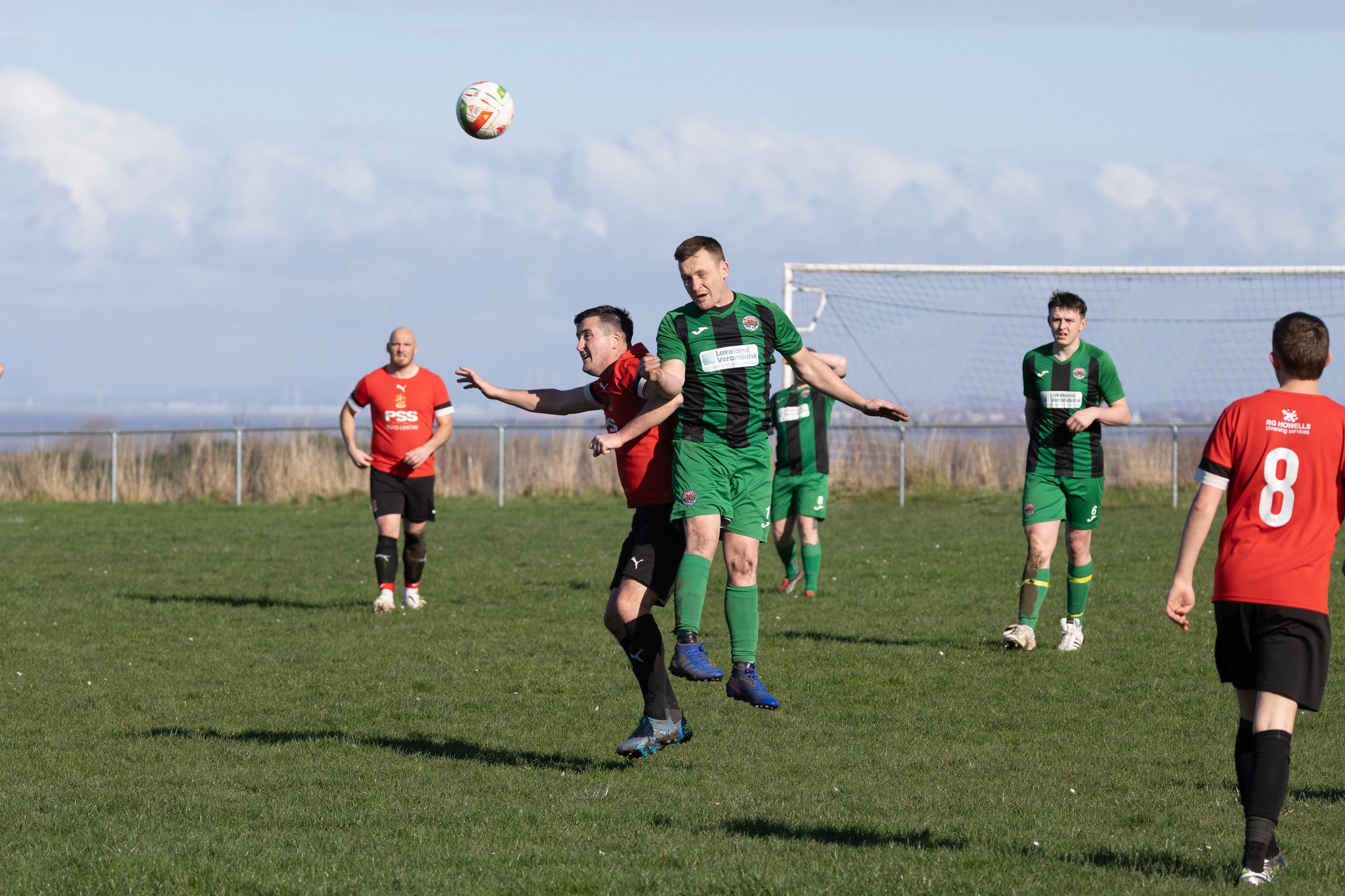 Soccer players in red and green jerseys contest a ball in midair on a grassy field with a goal in the background and blue sky with clouds overhead.