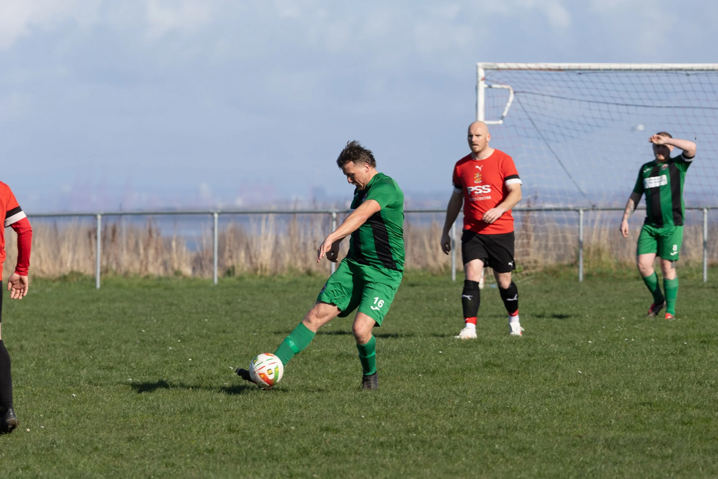 Soccer players on a grass field, one in green kicks a ball, others in red and black observe near goal net, under partly cloudy skies.