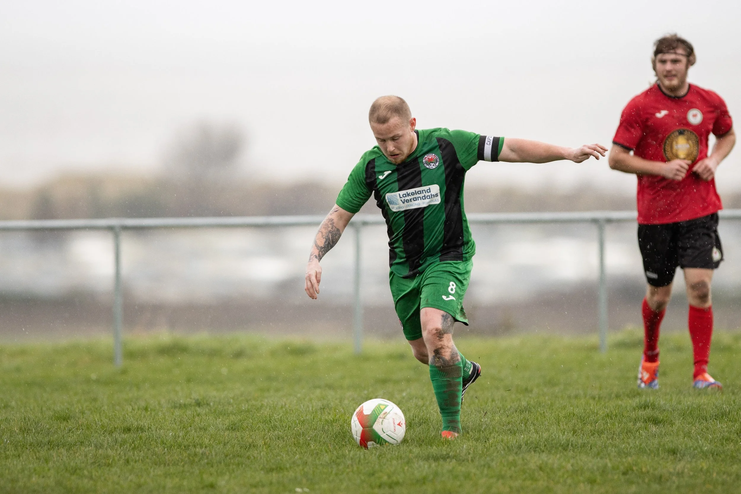 A soccer player in a green and black uniform is about to kick a ball on a grassy field while another player in a red uniform watches in the background.
