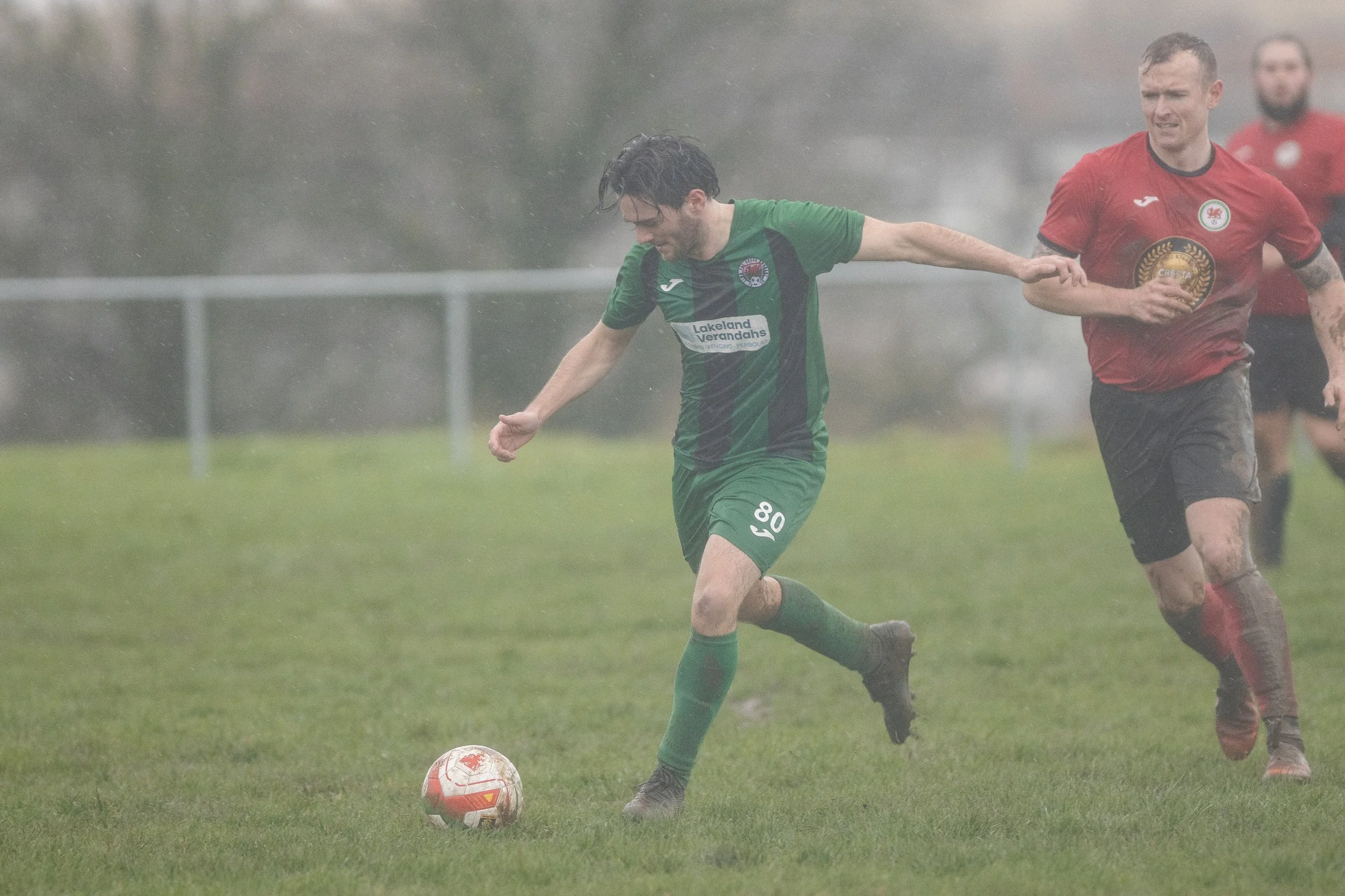 Soccer players playing in rainy weather on grass field, with one player in green jersey controlling the ball and others chasing.